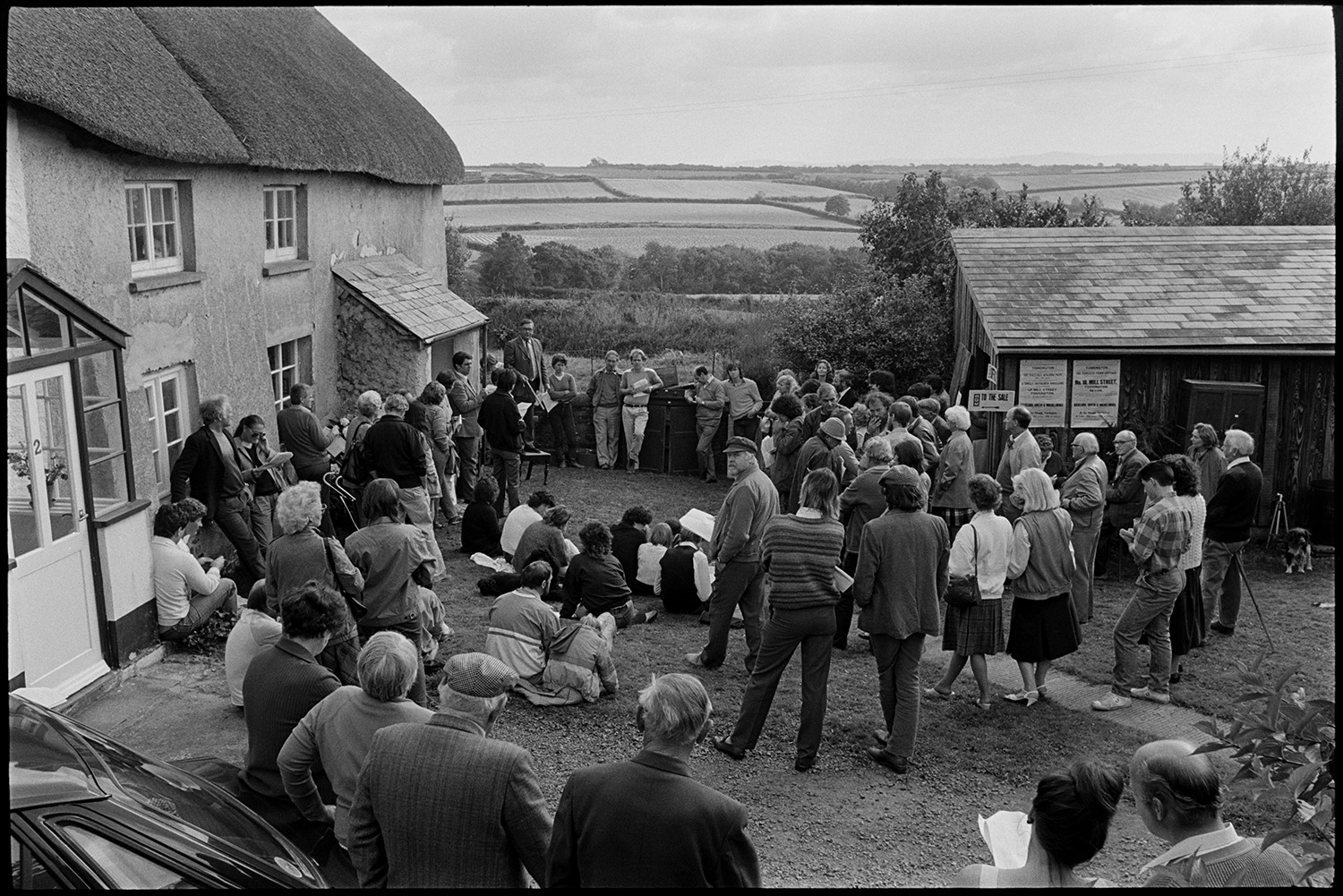 House auction, auctioneer standing on chair selling, man peering into house. Pot.
[A house auction at Roborough. Paul Foggo, the auctioneer, is stood on a chair by the doorway, with buyers and onlookers gathered around in the yard. A wooden shed is in the yard and fields are visible in the background.]