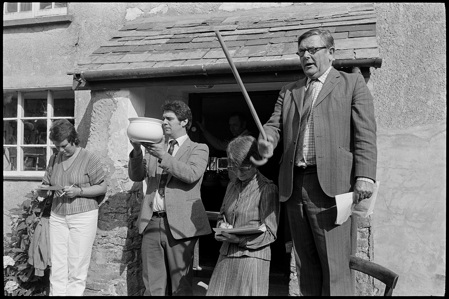 House auction, auctioneer standing on chair selling, man peering into house. Pot.
[A house auction at Roborough. Auctioneer Paul Foggo, is standing on a chair in front of the porch. He is holding up a walking stick and selling items. Diane Foggo is writing notes. Another man is holding up a chamber pot for sale.]