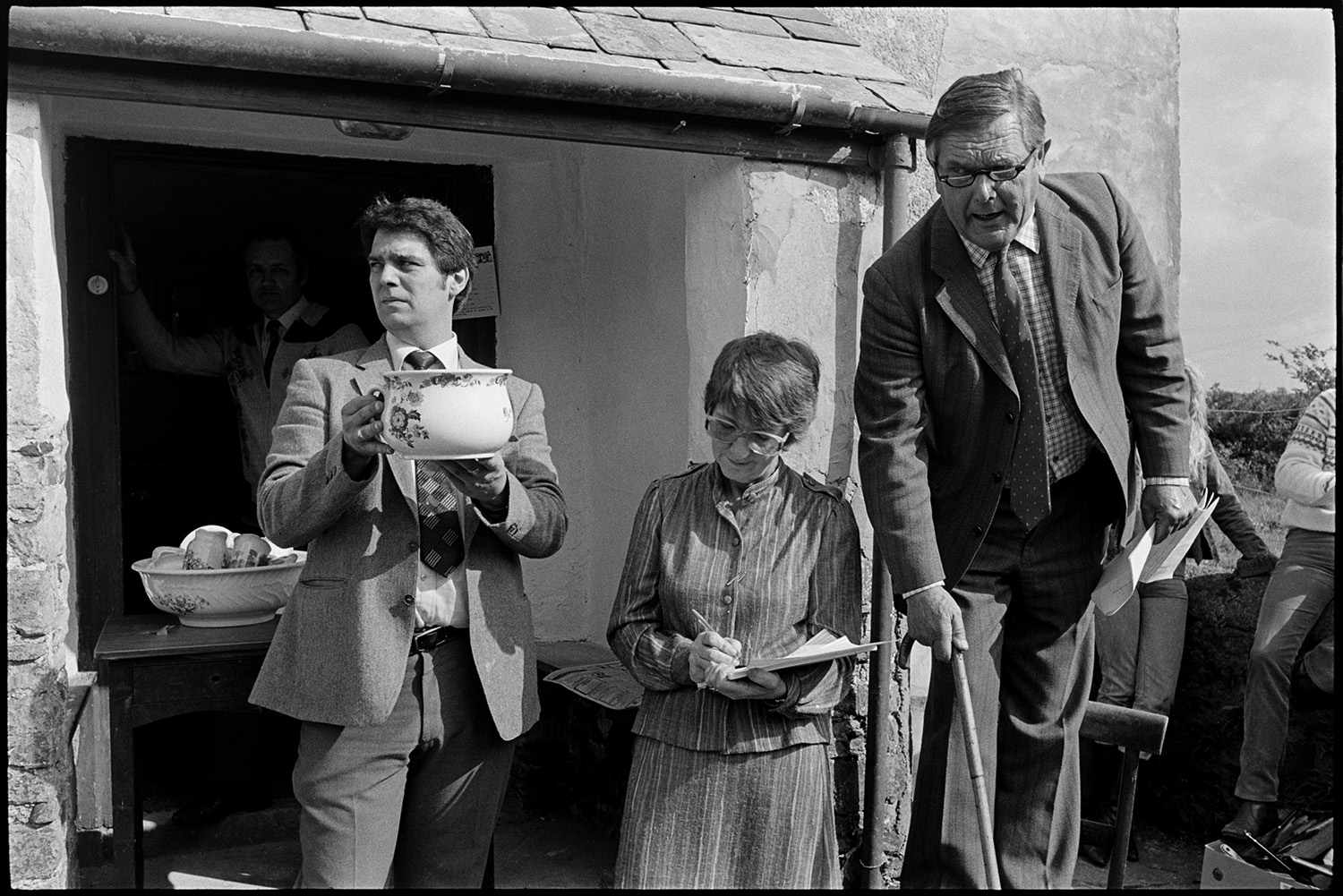 House auction, auctioneer standing on chair selling, man peering into house. Pot.
[A house auction at Roborough. Auctioneer Paul Foggo, is standing on a chair in front of the porch. He is holding a walking stick and selling items. Diane Foggo is writing notes. Another man is holding up a chamber pot for bids. A table with a bowl and china stands in the porch doorway.]