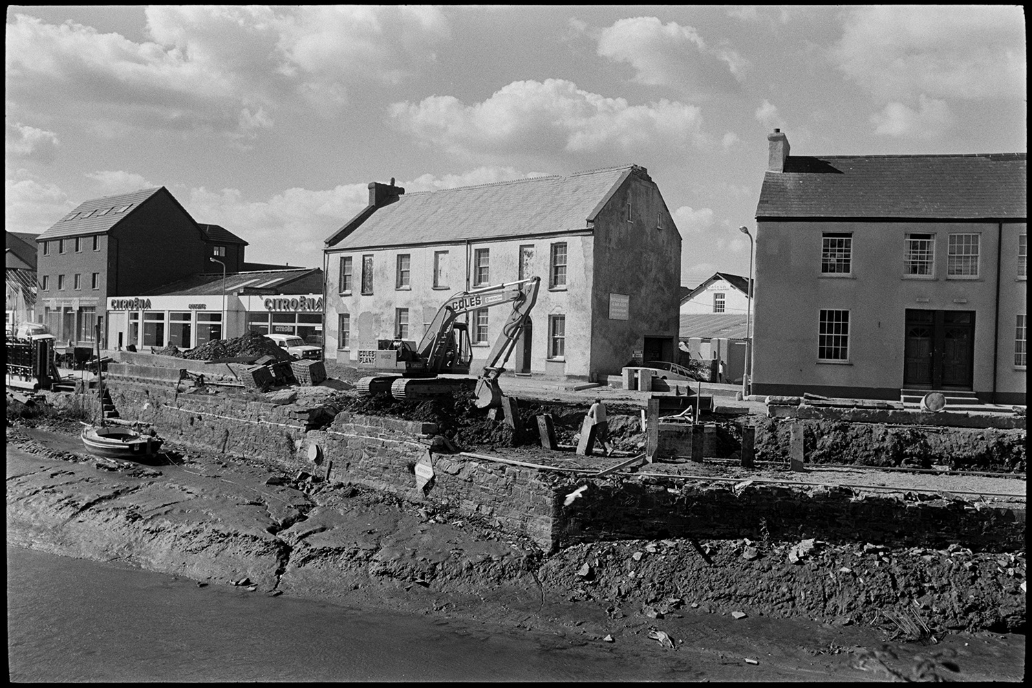 Repair work on quay, mechanical digger, quayside houses.
[Repair work, using a mechanical digger, to the quayside stone wall at Rolle Quay, Barnstaple. A small boat is on the mud bank. Old quayside properties stand beside a new car garage in the background.]