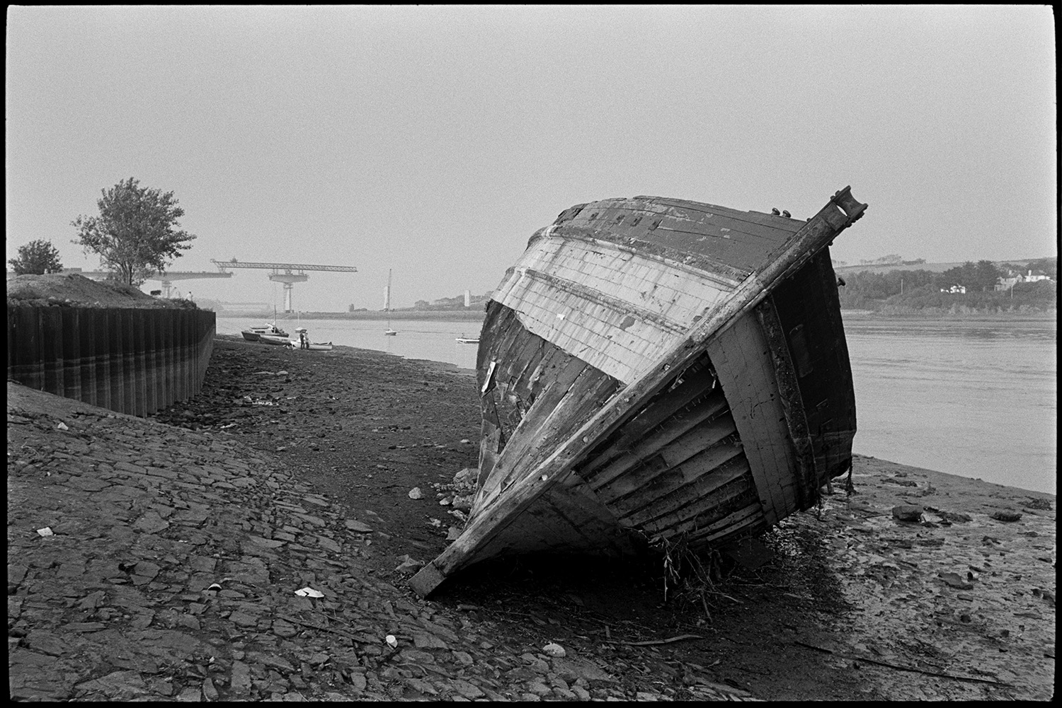 Remains of wooden fishing boat being broken up, new bridge construction behind.
[An old wooden fishing boat on the banks of the River Torridge at Bideford. Bideford New Bridge is being constructed in the background.]