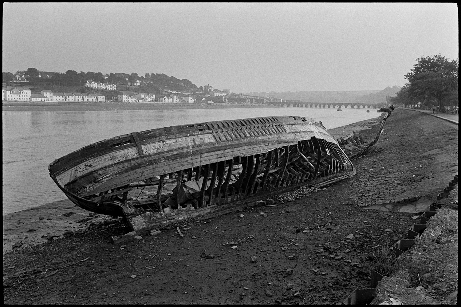 Remains of wooden fishing boat being broken up, new bridge construction behind.
[The remains of an old wooden fishing boat on the banks of the River Torridge. The boat is being broken up. Bideford and Bideford Long Bridge, also known as Bideford Old Bridge is visible in the background.]