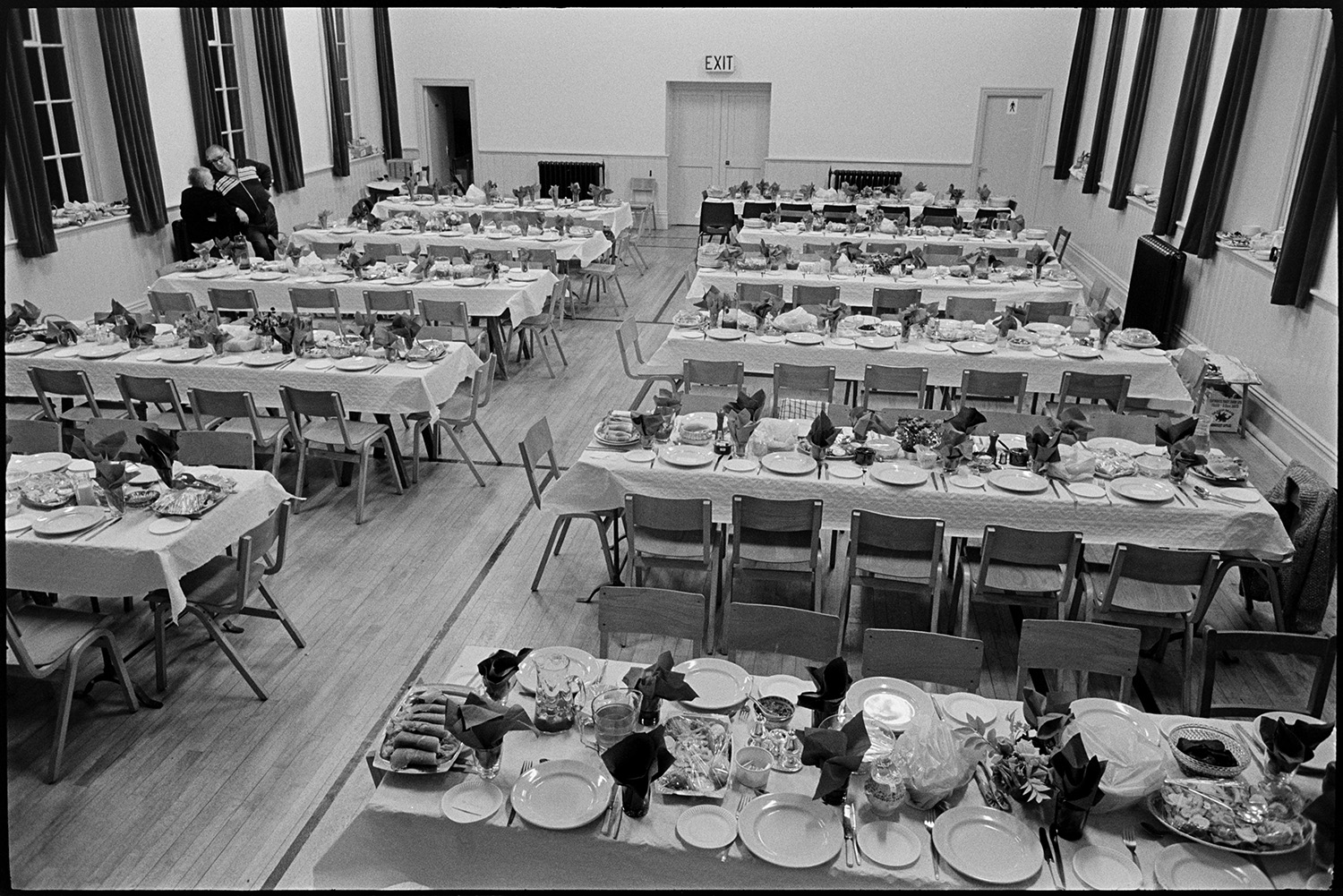 Women preparing supper, dinner in village hall, details of place settings, china, cutlery.
[Tables laid in preparation for a dinner at Winkleigh Village Hall, possibly an Over 60s Club dinner. China, cutlery, glasses, and some covered plates of food are on the tables. A man and woman are talking at the side of the hall.]