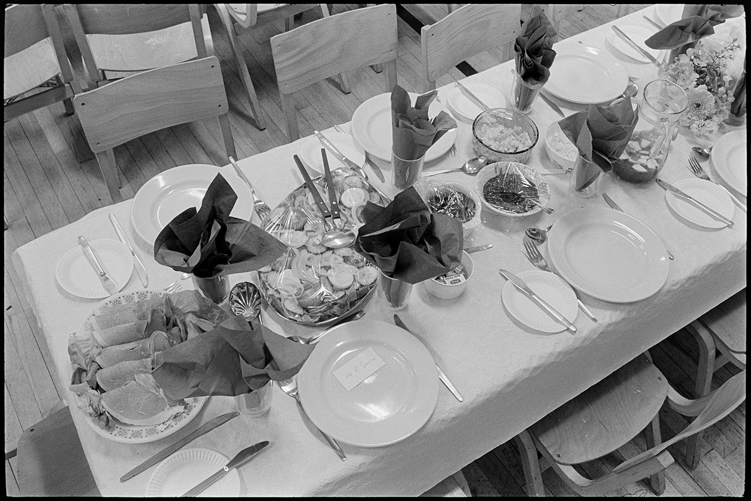 Women preparing supper, dinner in village hall, details of place settings, china, cutlery.
[A table laid for dinner at Winkleigh Village Hall, possibly for an Over 60s Club dinner. China, glasses with serviettes, cutlery, and some covered plates of food, including cold meats, are visible.]