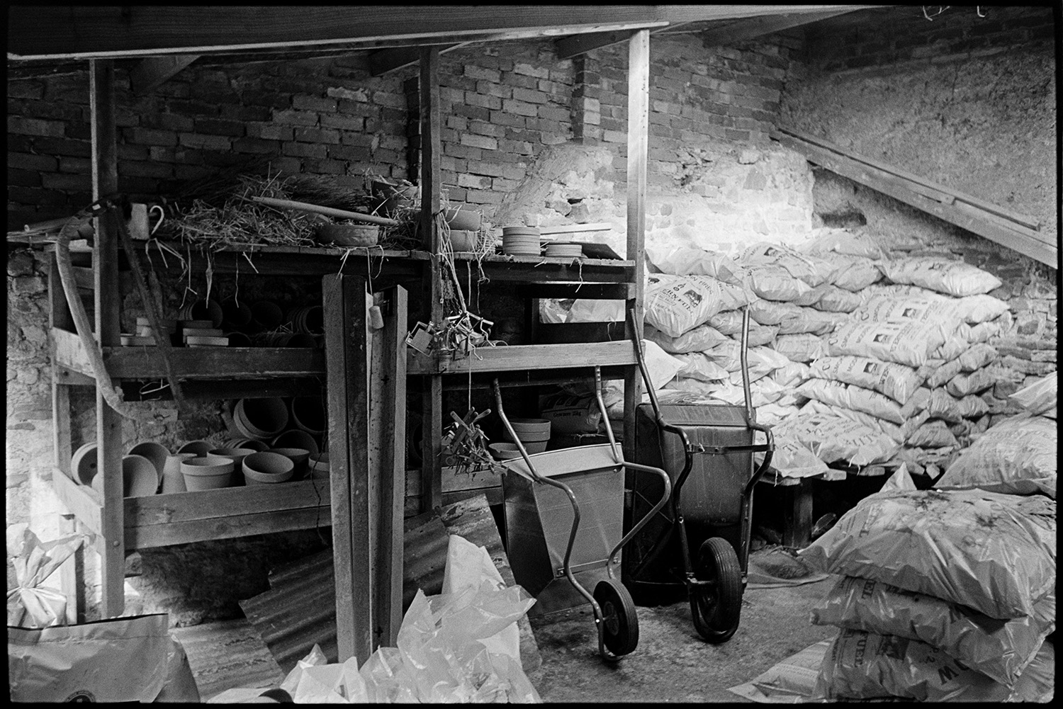 Interior of ironmongers shop, shelves with china, coal store. Proprietors at counter.
[Interior of the coal store at Ellacott's ironmongery in Market Street, Hatherleigh. There are two wheelbarrows, wooden shelving with plant pots and garden goods, alongside a stack of plastic sacks of coal.]