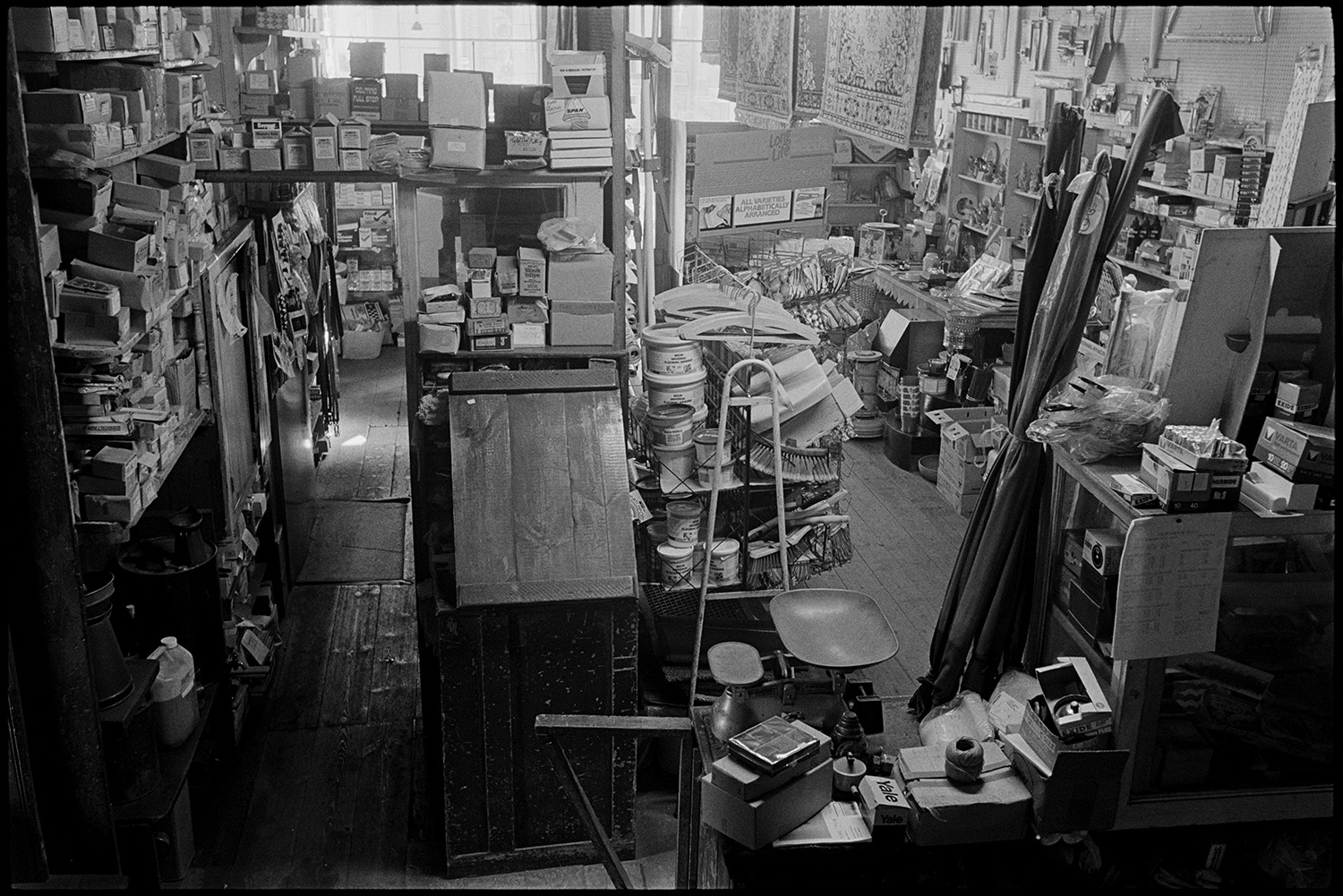 Interior of ironmongers shop, shelves with china, coal store. Proprietors at counter.
[Interior of Ellacott's ironmongery in Market Street, Hatherleigh. A wooden counter is shown with weighing scales, and is surrounded by shelves and racks full of various goods.]