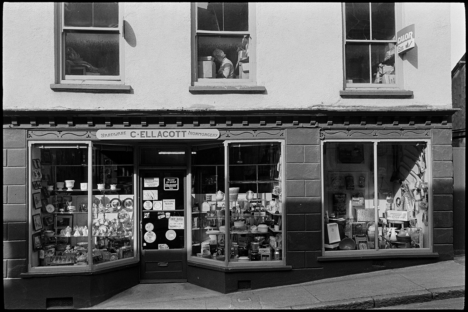 Front of ironmonger's shop. Exterior.
[Exterior of Ellacott's ironmongery shop in Market Street, Hatherleigh. China ornaments, pots and kitchen goods are on display in the shop front window. A woman is shown working in a upstairs window. There is a Calor gas sign, and a handwritten shop sign on the building.]