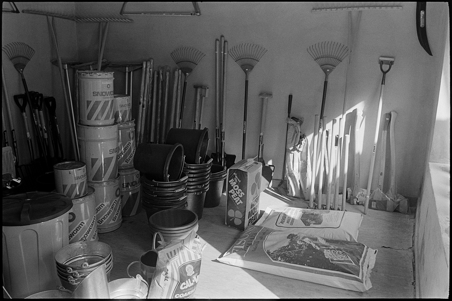 Interior of ironmongers shop, shelves with china, coal store. Proprietors at counter.
[Interior of the store room at Ellacott's ironmongery shop in Market Street, Hatherleigh. Garden tools are propped against the wall, with paint tins, buckets, and plastic bags of compost on the floor.]