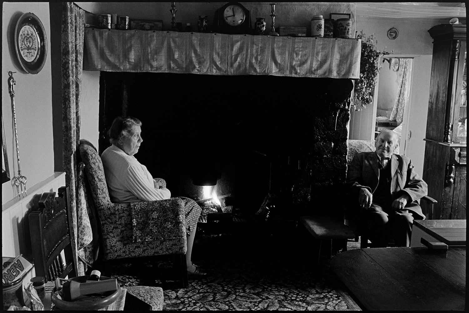 Cottage interior. Man and woman seated by open fire, reading, dresser with china, side table.
[Edith Crocker and Henry Crocker, brother and sister, are sitting by an open fire in their cottage in Monkokehampton. They are both sitting in armchairs. A royal wedding plate and toasting fork hang on the wall and a clock, candlesticks and jars are displayed on the mantelpiece above the fireplace.]
