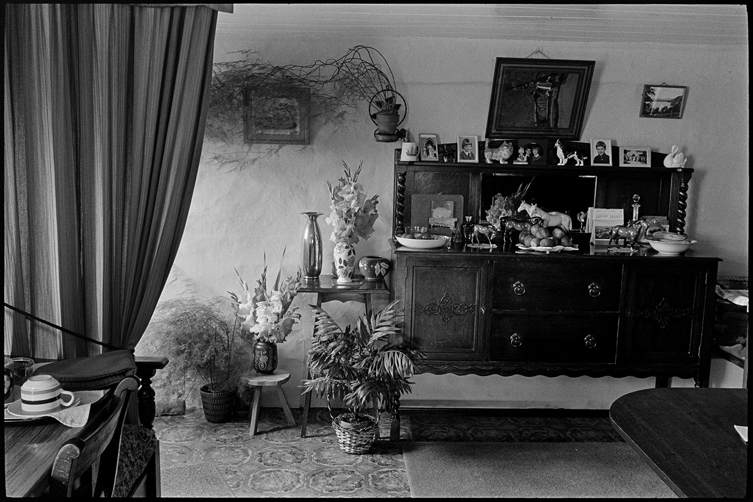 Cottage interior. Man and woman seated by open fire reading, dresser with china, side table.
[The interior of Edith Crocker and Henry Crocker's cottage at Monkokehampton. A large wooden sideboard with china ornaments, fruit bowls and photographs on it is visible. There are pictures on the wall, with pot plants and flowers on display around the sideboard.]