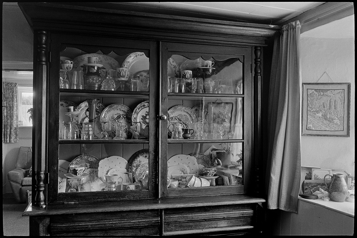 Cottage interior. Man and woman seated by open fire reading, dresser with china, side table.
[The interior of Edith Crocker and Henry Crocker's cottage at Monkokehampton, showing a large dresser with glass doors. It is full of china, plates, crockery and glassware. There is a curtain and window ledge beside it, and another window and armchair in the room behind.]