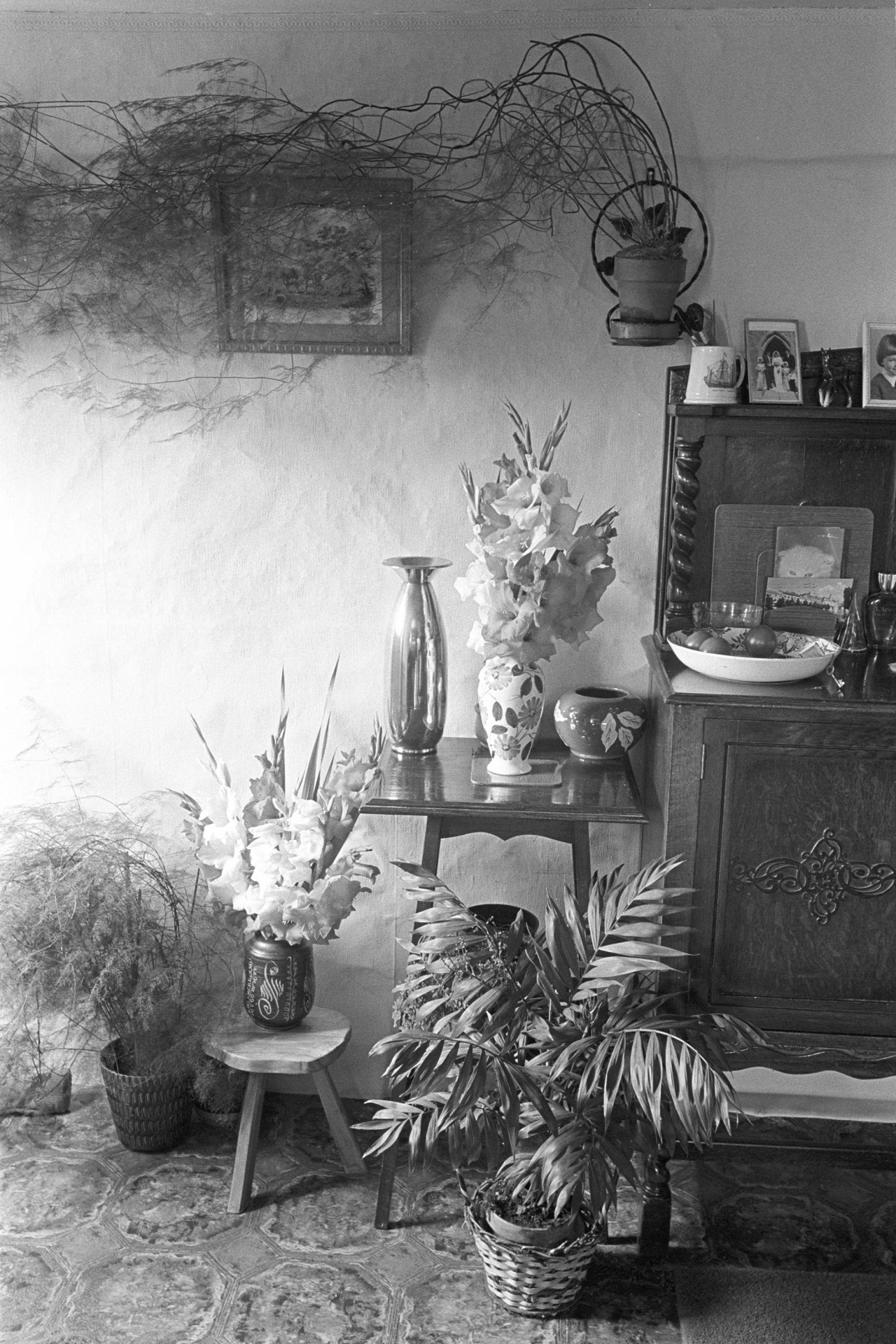 Cottage interior, side table with ornaments and flowers. 
[The interior of a cottage in Monkokehampton with a wooden side table and various pot plants and flowers in vases. One pot plant is mounted on the wall by a framed picture.]