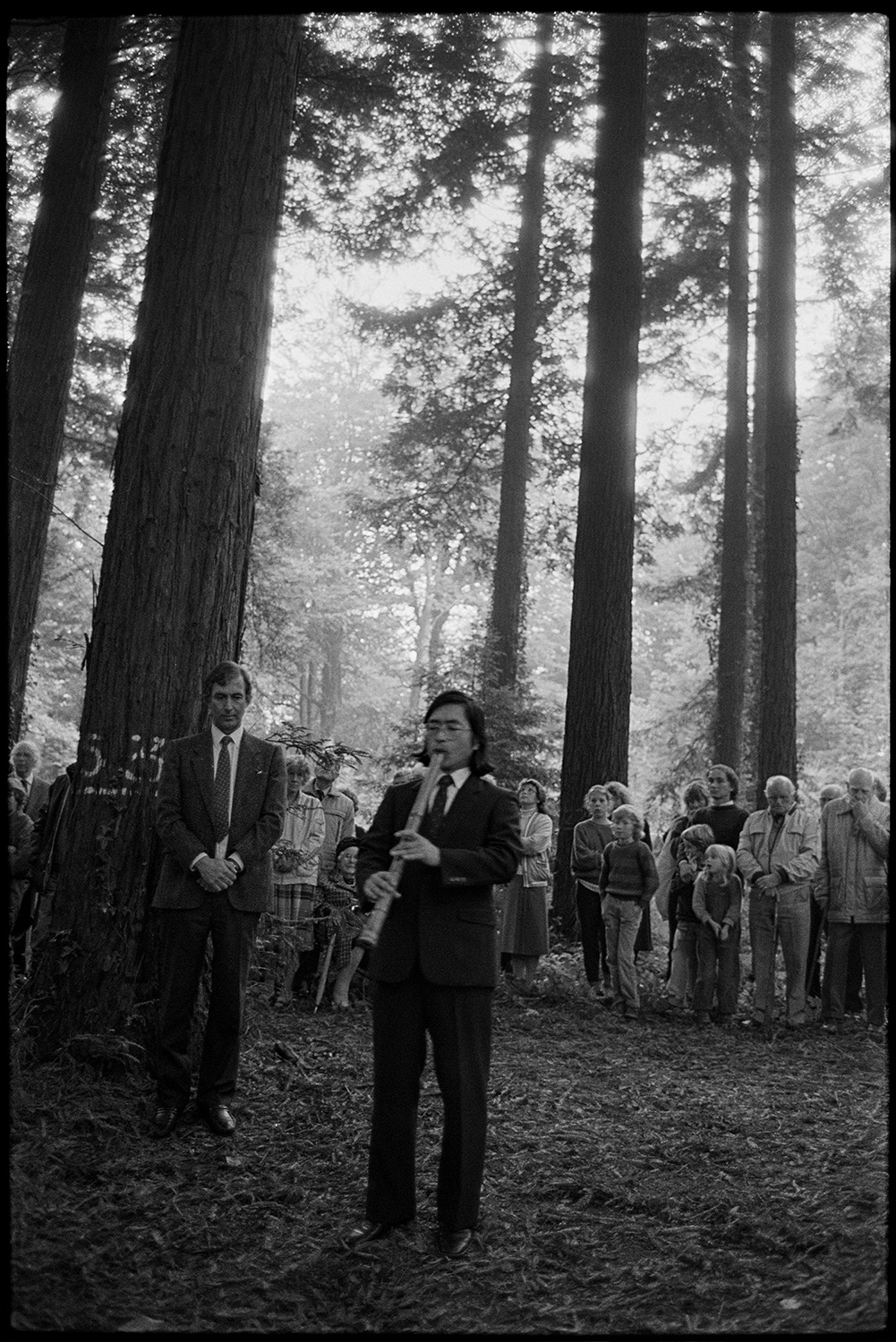 Spectators watching and listening to a musician playing a woodwind instrument at a ceremony in Dartington. The musician is wearing a dark suit and tie. There are tall fir trees in the background.