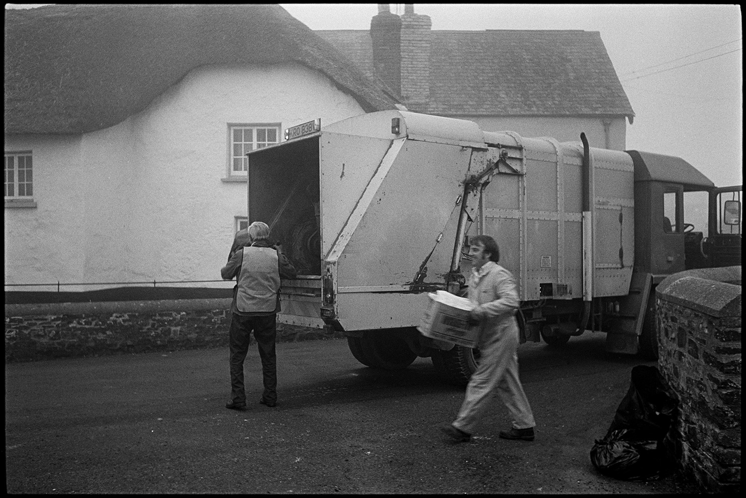 Dustcart lorry in street with men loading rubbish bags.
[A dustcart lorry in the street at Dolton with two men loading bin bags and rubbish into it. A thatched cottage is in the background.]
