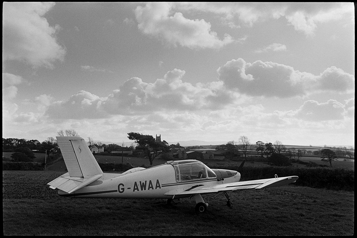 Light aeroplane parked in field, belonged to farmer.
[A light aeroplane, belonging to Mr Dunn, parked in a field at Dowland. Dowland church tower is visible in the background, with farm buildings and houses. Dartmoor is also visible in the far distance, under a cloudy sky.]