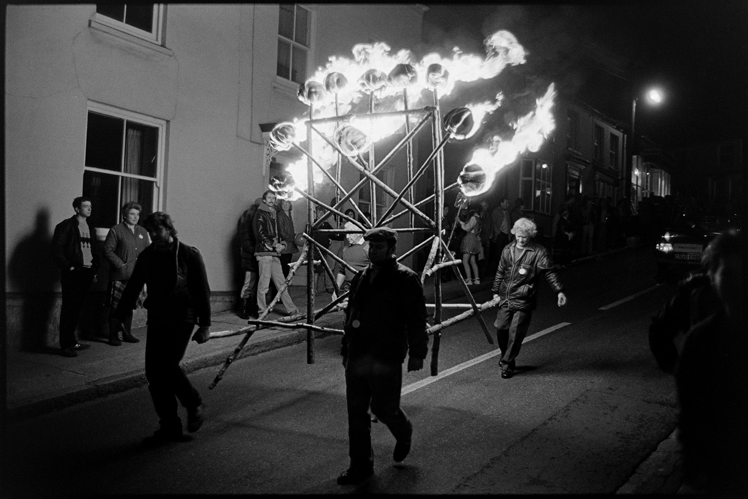 Carnival procession at night, with burning torches and blazing tar barrels, floats.
[Four men walking down a street carrying a timber frame of burning torches at Hatherleigh Carnival. Spectators are lining the street watching.]