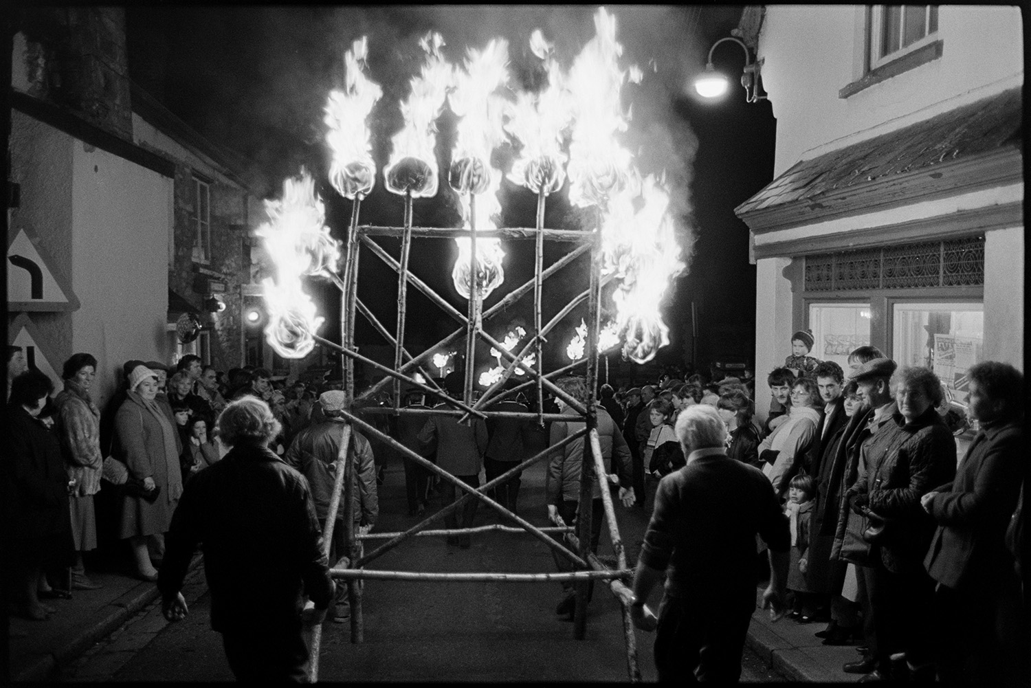 Carnival procession at night with burning torches and blazing tar barrels, floats.
[Four men walking down a street carrying a timber frame of burning torches  at Hatherleigh Carnival. Spectators are lining the street watching.]
