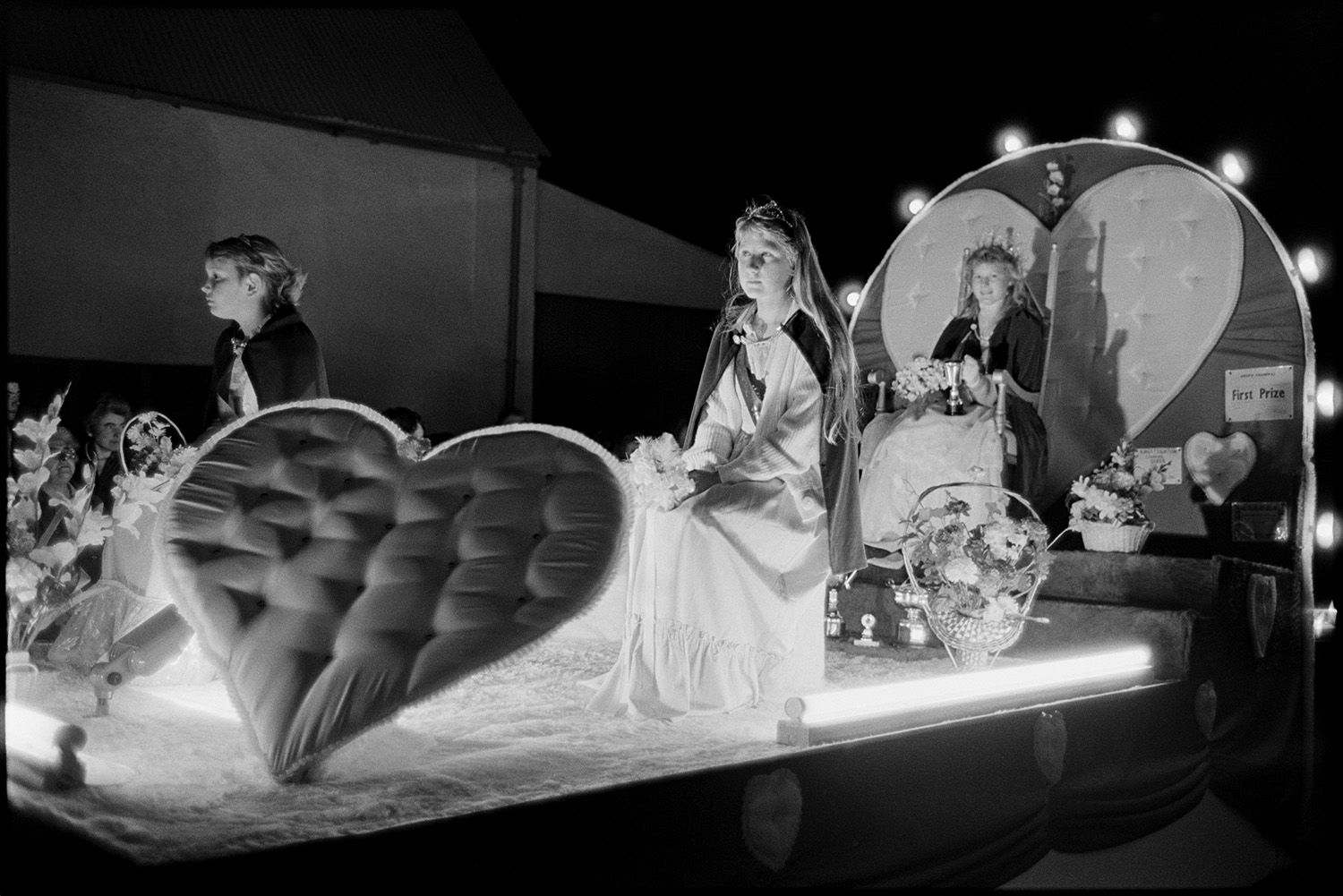 Carnival procession at night. Queen and attendants and other floodlit floats with children.
[Three girls sitting on a carnival float decorated with hearts and flowers during the evening procession at Hatherleigh Carnival. There is a first prize certificate and trophies on display.]