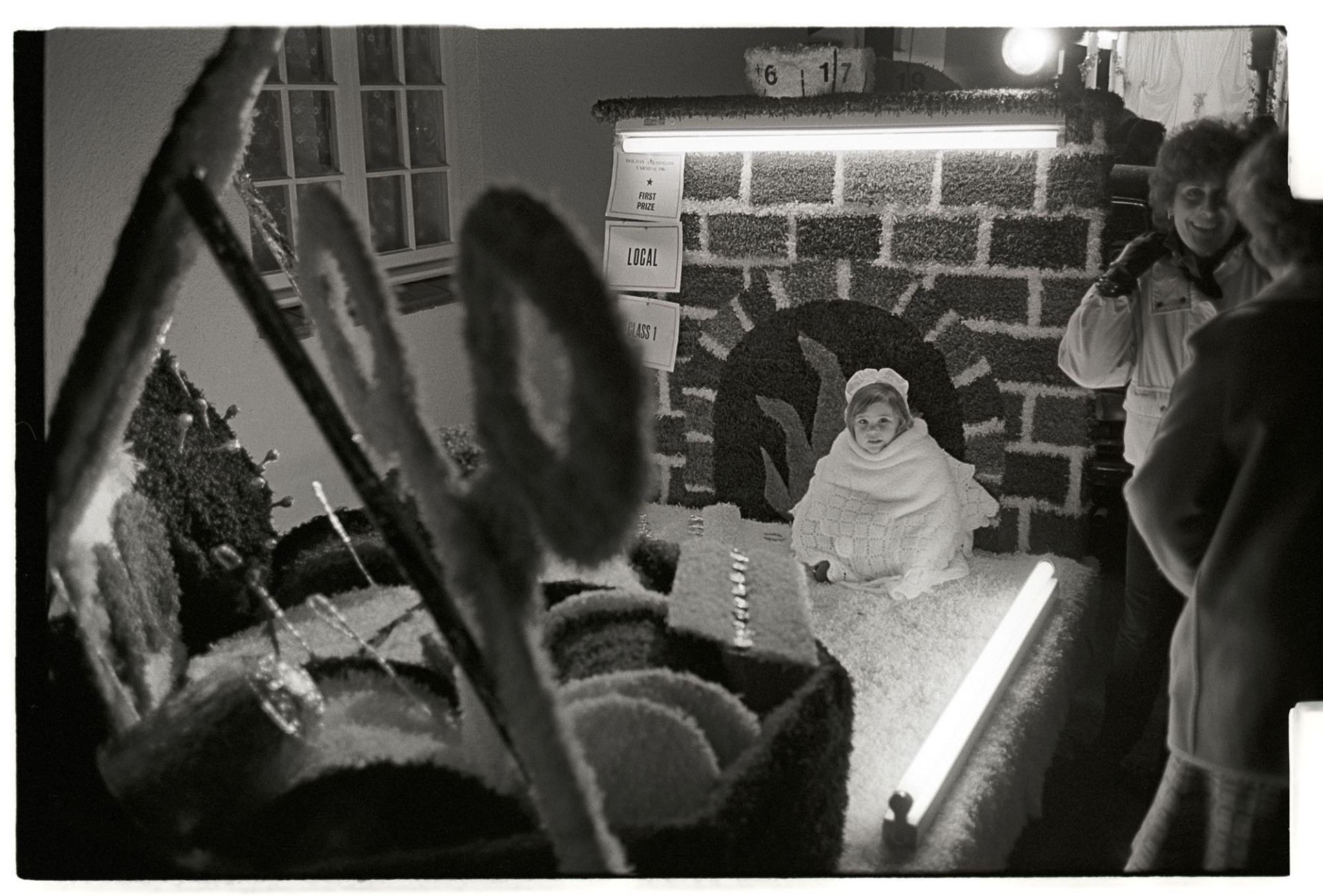 Carnival at night, child on lit float in fancy dress. 
[A child in fancy dress on a carnival float at Dolton carnival at night. The child is sat by the model of a fireplace. Two spectators are talking by the float.]