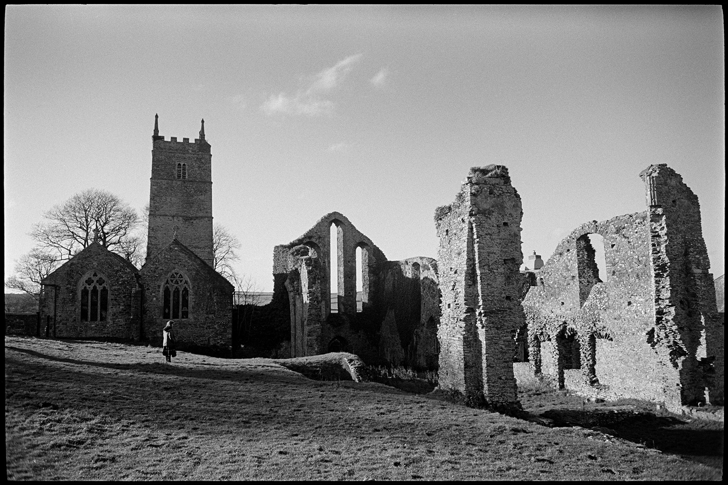 Ruined priory and graveyard.
[The ruined remains of Frithelstock Priory, next to the Church of St Mary and St Gregory, Frithelstock. A woman is standing and looking at the ruins.]