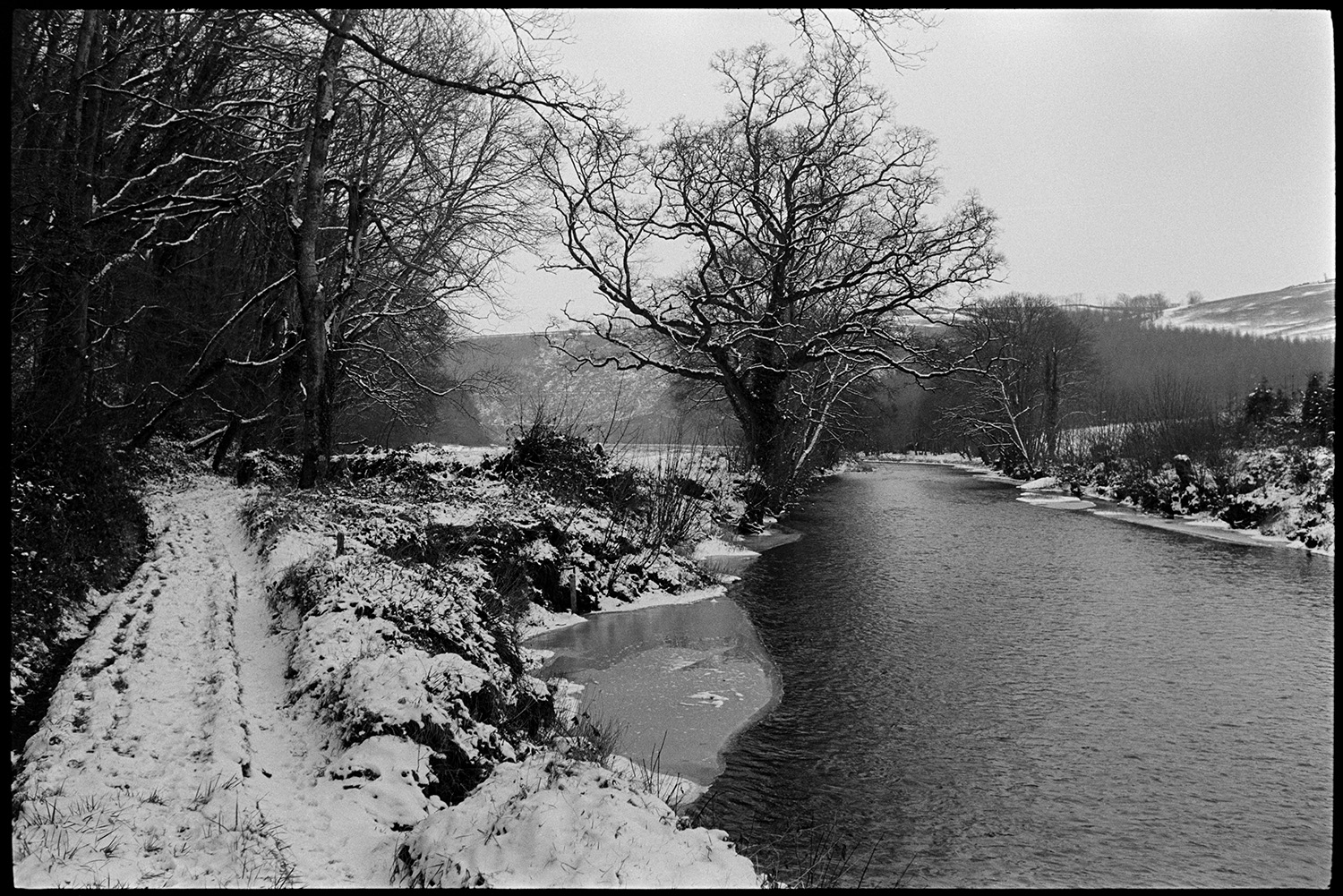 Snow, river and wood.
[River Torridge in winter with snowy banks and ice, and view of trees lining the river, at Halsdon, Dolton. A snow covered track is running alongside the river.].