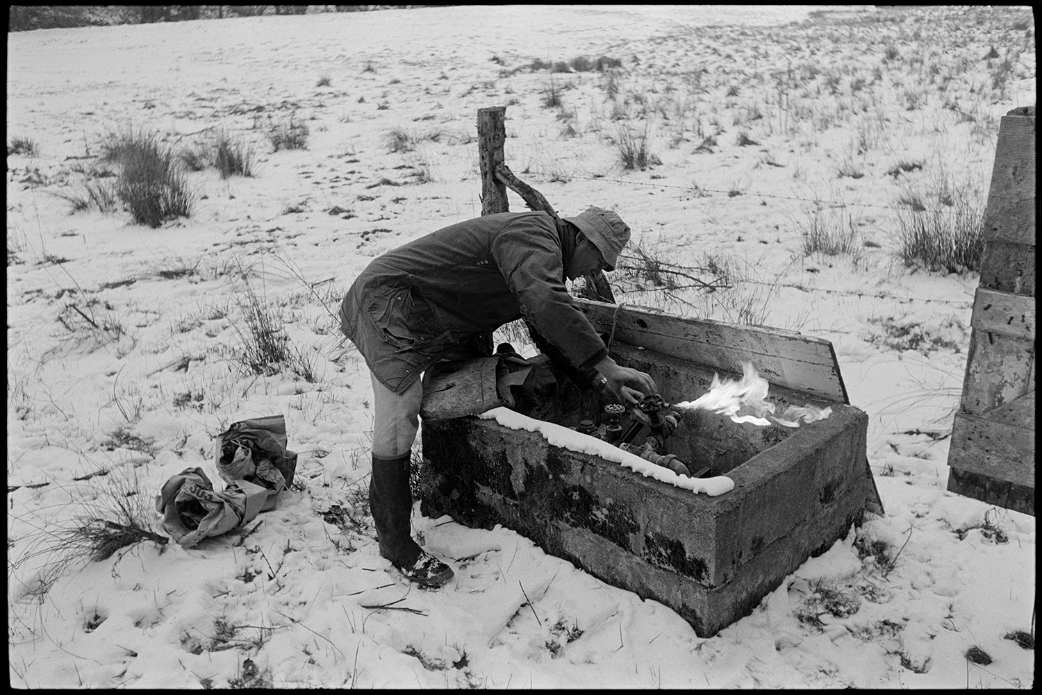 Snow, farmer and dogs breaking ice and trying to start tractor with frozen diesel fuel.
[Guy Crossman defrosting a frozen pump in a field covered in snow at Great Warham, Beaford.]