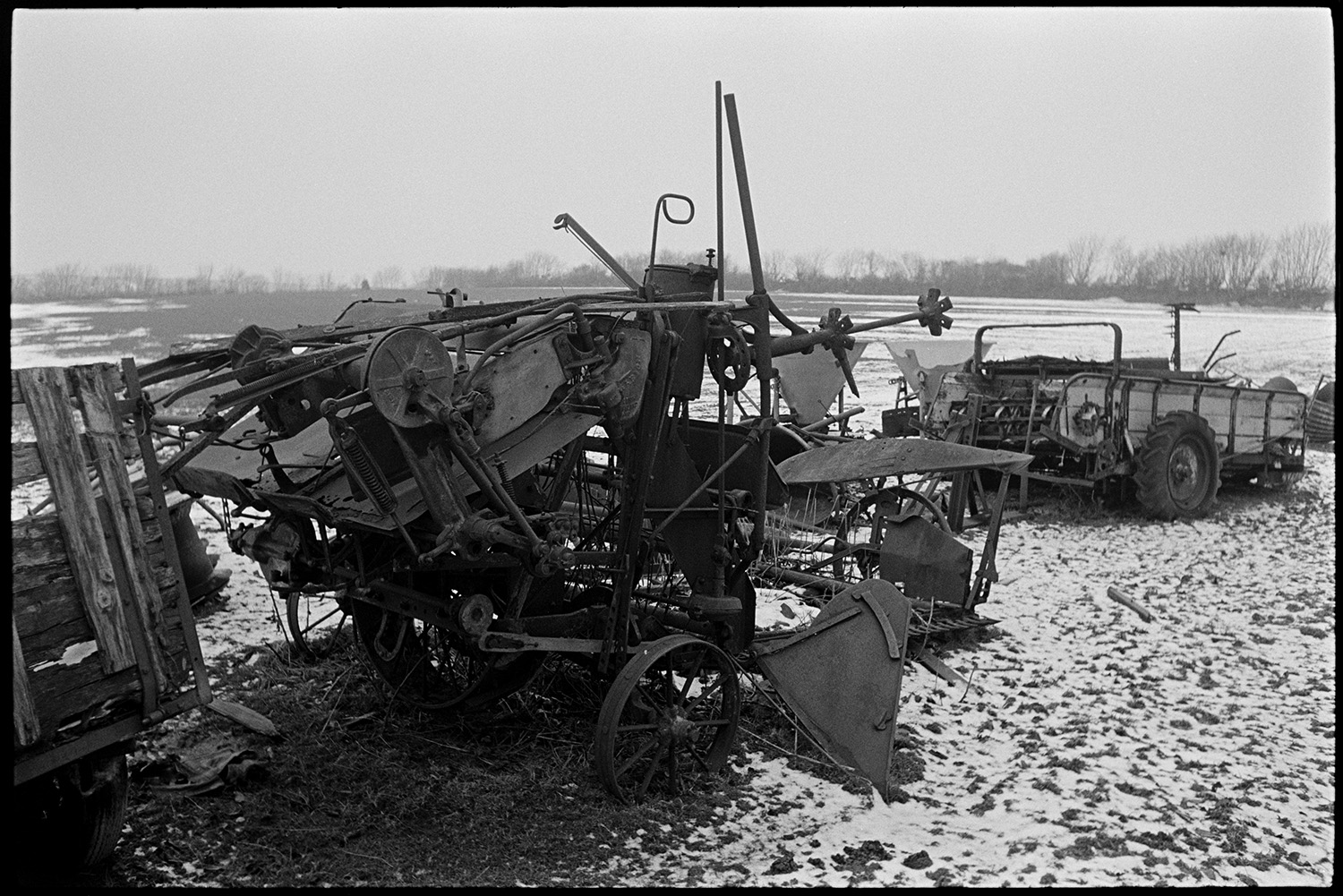 Snow, old rusting implements lying in field, plough, circular saw, reap and binder.
[Old plough and other disused farm equipment piled in a field lightly covered in snow, at Ashwell Farm, Dolton.]