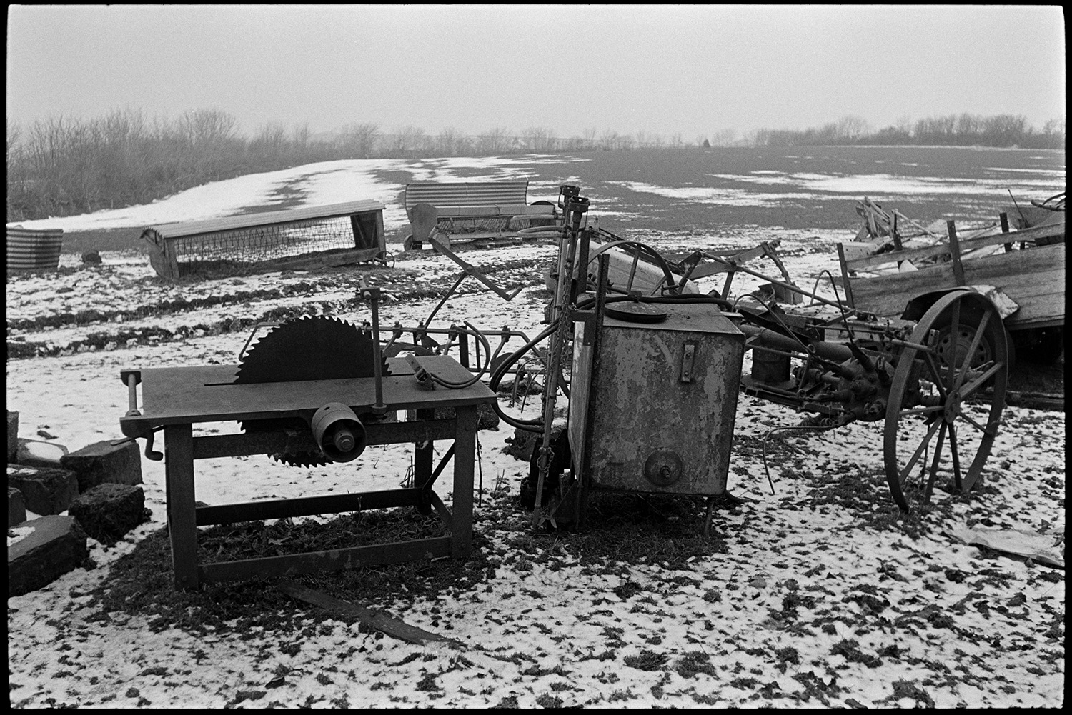 Snow, old rusting implements lying in field, plough, circular saw, reap and binder.
[Circular saw bench, pieces of old farming equipment and hay feeders in a field lightly covered with snow at Ashwell Farm, Dolton.]