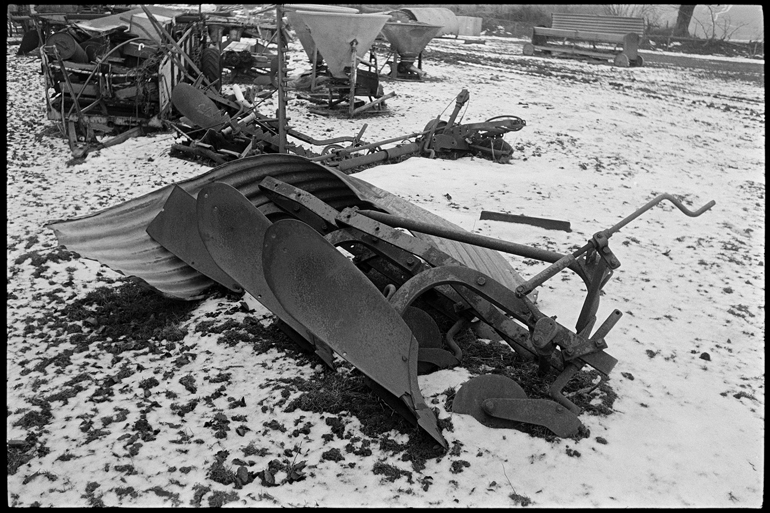 Snow, old rusting implements lying in field, plough, circular saw, reap and binder.
[Old plough, corrugated iron, hay feeders and other disused farm equipment piled in a field lightly covered with snow at Ashwell Farm, Dolton.]