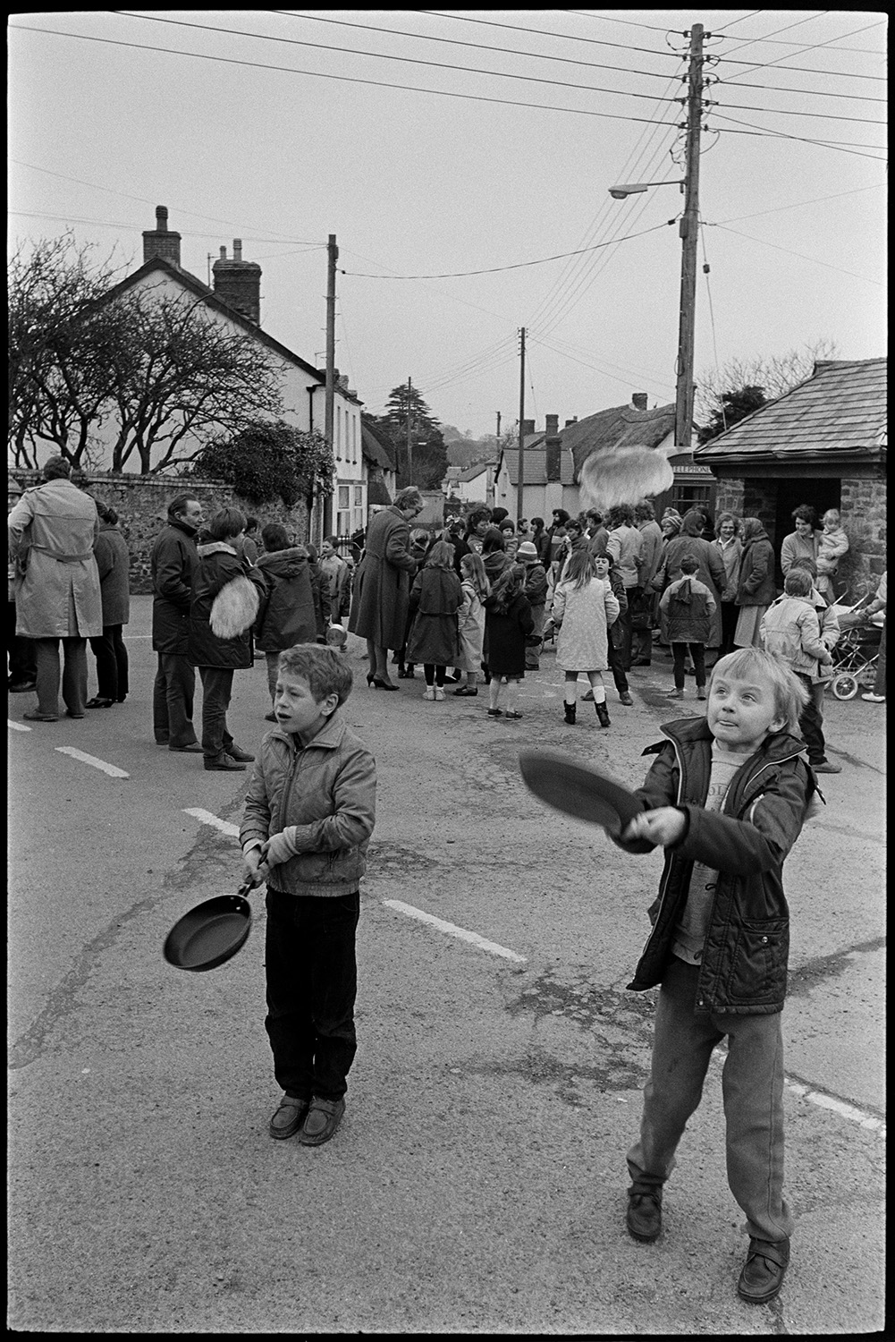 Shrove Tuesday Pancake Race in village street, children's races.
[Two boys holding frying pans and tossing pancakes into the air in Fore Street, Dolton in a Shrove Tuesday Pancake Race. In the background there is a crowd of men, women and children in the road and a view of thatched houses.]