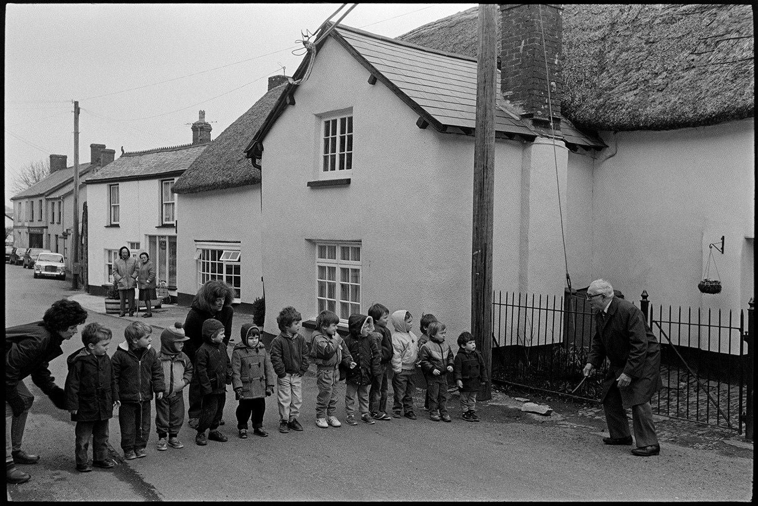 Shrove Tuesday Pancake Race in village street, children's races.
[Thirteen young children lined up across Fore Street, Dolton, dressed in winter clothes, getting ready for a race in the Shrove Tuesday pancake day celebrations. A man and two women are starting the race. In the background a man and woman are stood outside the Post Office watching.]