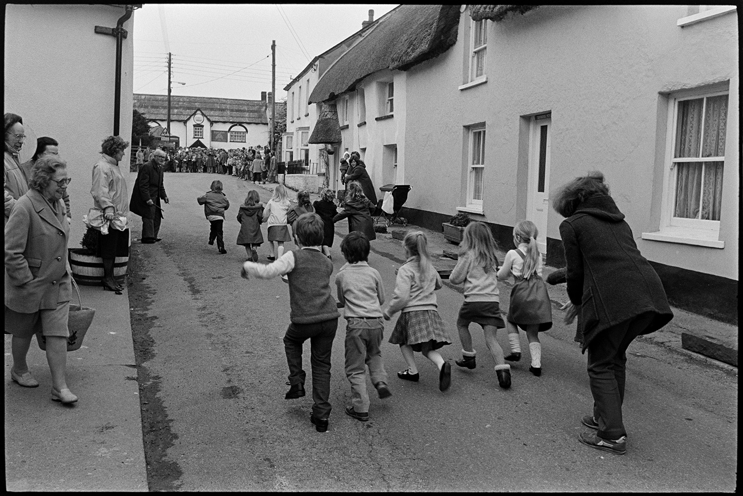 Shrove Tuesday Pancake Race in village street, children's races.
[Eleven children holding frying pans with pancakes in a Shrove Tuesday Pancake Race along Fore Street, Dolton, with a crowd of men, women and children watching. In the background is a view of the front of the Royal Oak pub and thatched houses.]