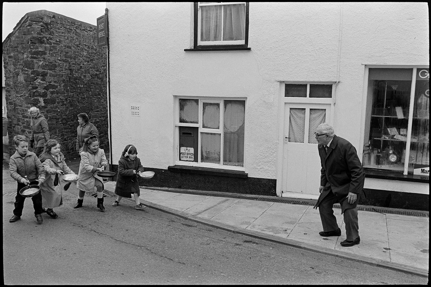 Shrove Tuesday Pancake Race in village street, children's races.
[Four children holding frying pans containing pancakes in a Shrove Tuesday Pancake Race along Fore Street, Dolton. They are being watched and encouraged by a man and two women as they pass the Post Office.]