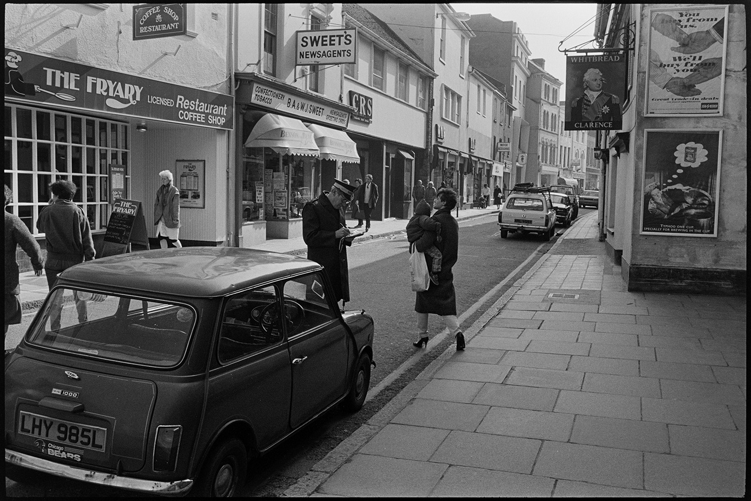 Street scene in High Street, Barnstaple, showing a traffic warden completing a parking ticket for a car parked on a restricted parking area.  Pedestrians are walking along pavements and shop signs can be seen for The Fryary restaurant, B.A. & W.J. Newsagents shop, and the Clarence pub which is now called the Cork and Bottle.