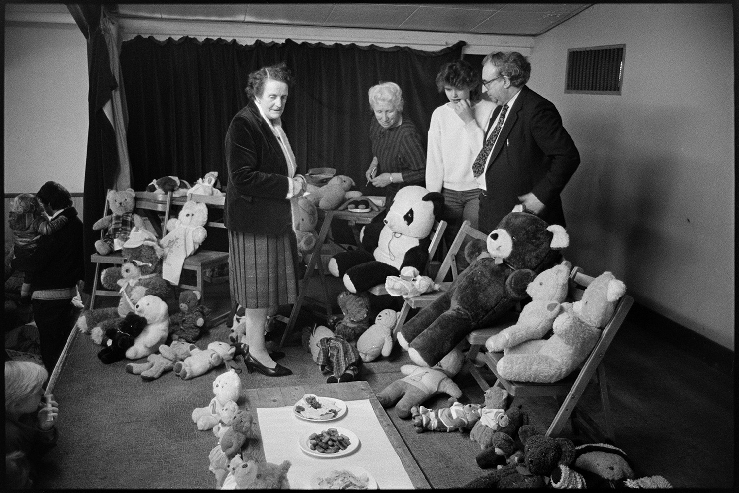 Easter show in village hall teddy bears party, competition for the oldest bear.
[Three women and a man looking at a collection of teddy bears which have been entered into a Teddy Bears Party Competition for the oldest bear, in the Easter Show at Dolton Village Hall.]