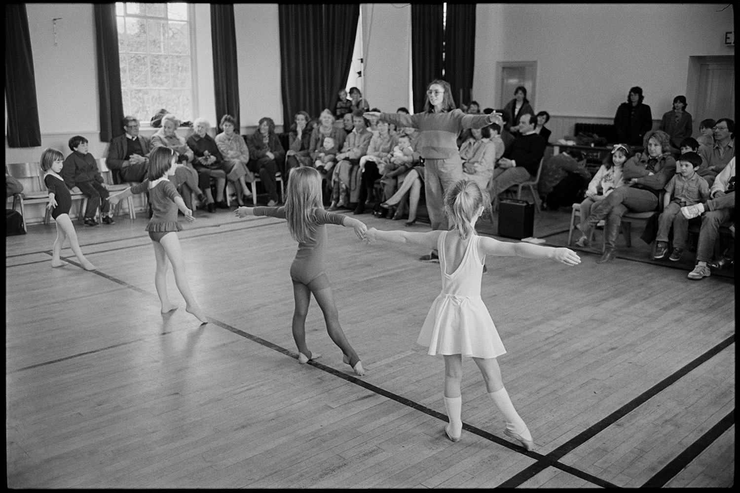 Dance class, children dancing performance in front of parents. Sally Barber's dancing.
[Four girls dancing in Winkleigh Village Hall in a performance by Sally Barber's dancing class in front of their parents, with Sally Barber standing in front of the girls directing them.]