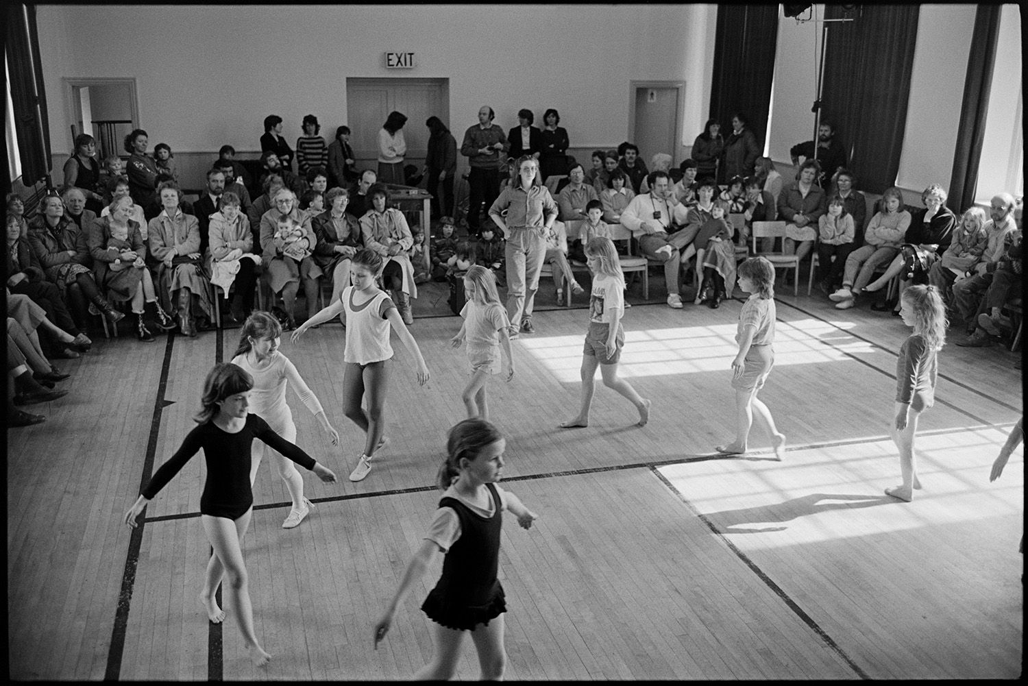 Dance class, children dancing performance in front of parents.
[A group of girls dancing in a circle at Winkleigh Village Hall in a performance by Sally Barber's dance class in front of parents and family, with Sally Barber directing the dancing.]
