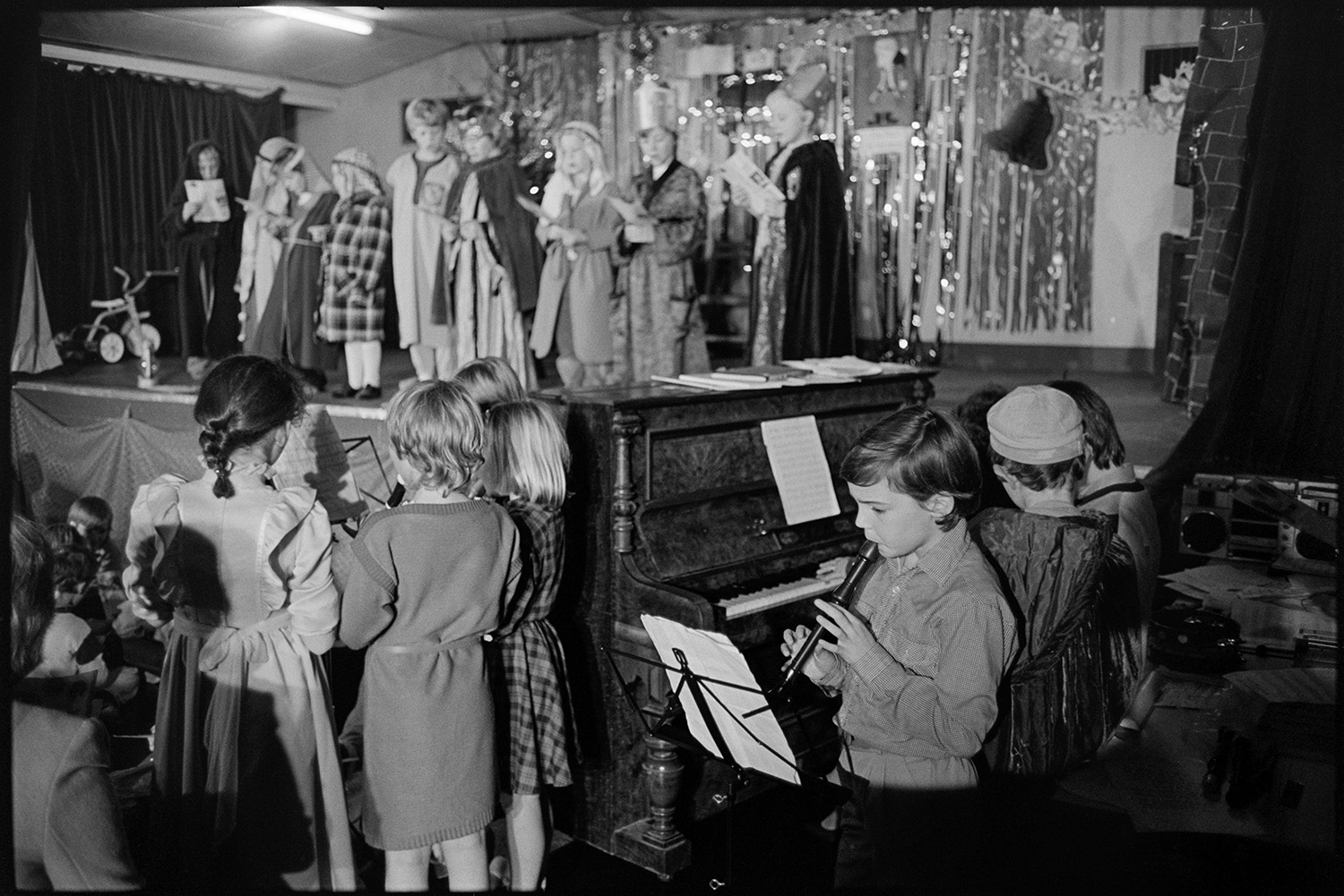 Christmas show in village hall Nativity play, children performing.
[Children singing in a Christmas show on the stage in Dolton Village Hall, dressed as characters in the Nativity story. Other children accompanying them on recorders and a piano in the foreground.]