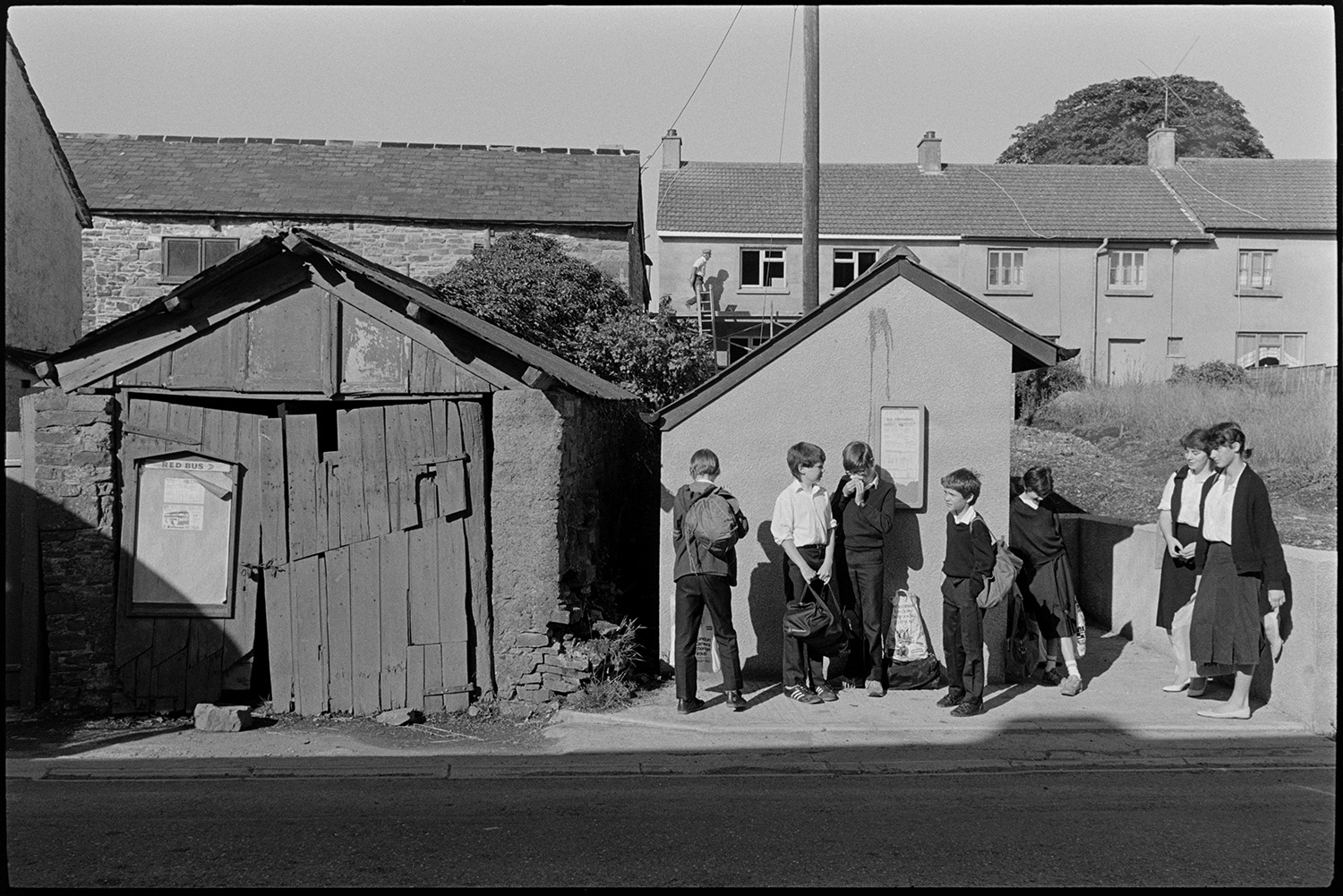 School children waiting for bus in village street beside old shed, garage.
[Seven children waiting for a bus, standing by the bus shelter in Beaford. Houses and gardens are visible in the background. A man working on top of the porch of one of the houses is also visible. Next to the bus shelter is a garage with an old wooden door on which is fixed a Red Bus noticeboard.]