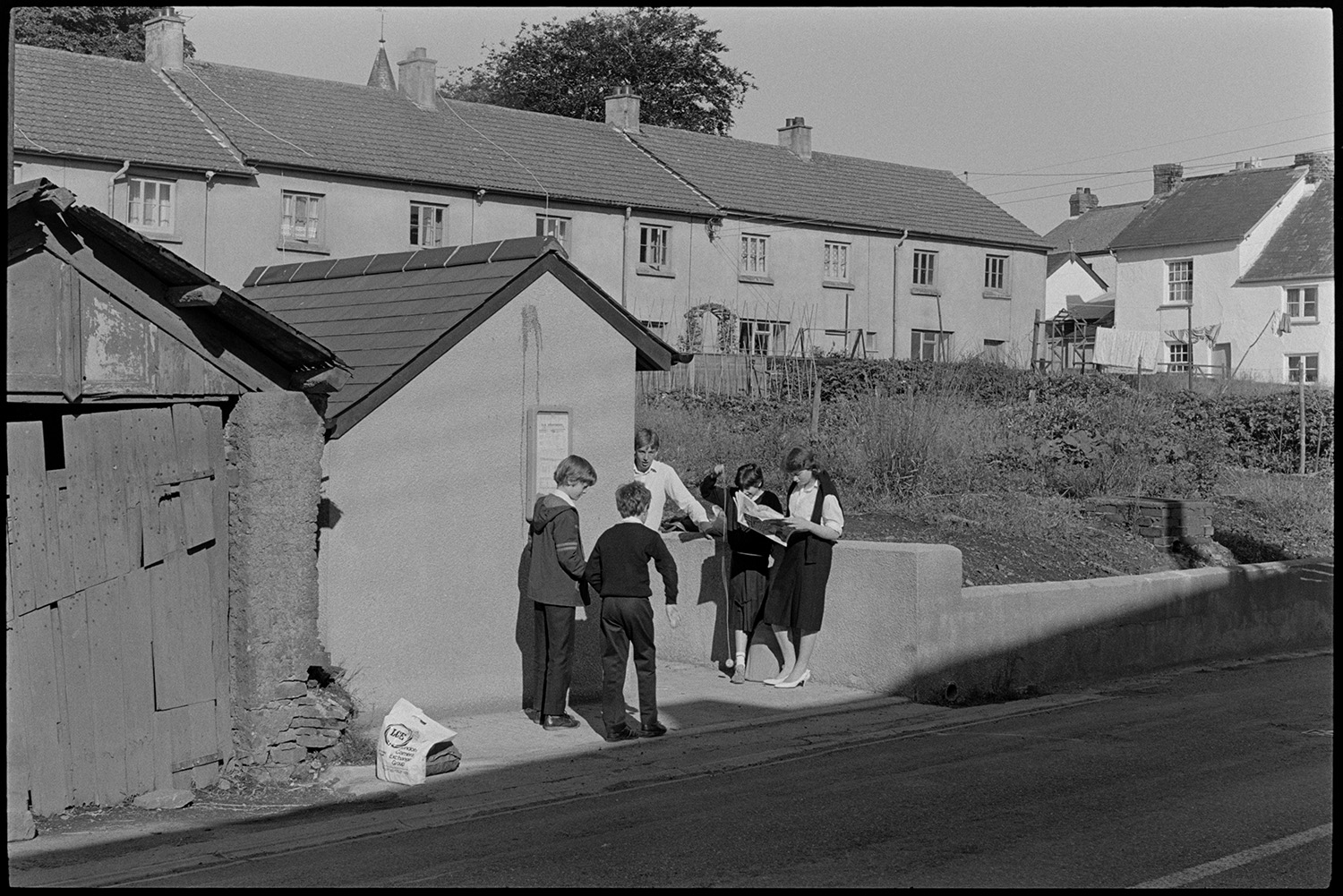 School children waiting for bus in village street beside old shed, garage.
[Five children waiting for a bus, standing by the bus shelter in Beaford. One girl is playing with a yoyo. Houses and gardens are visible in the background and next to the bus shelter is a garage with an old wooden door.]