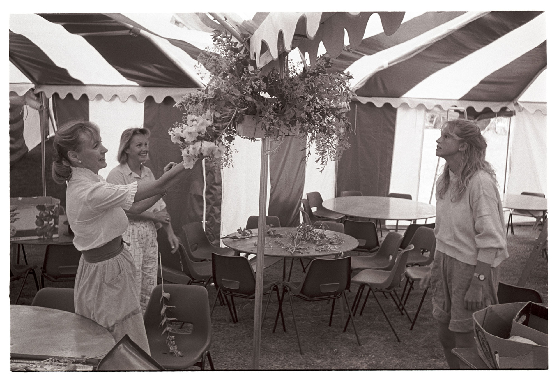 Women decorating marquee tent for hunt ball with flowers. 
[Lady Stukley, standing on the left, and two other women arranging a flower display in a marquee at Colleton Manor, Chulmleigh, for a hunt ball.]