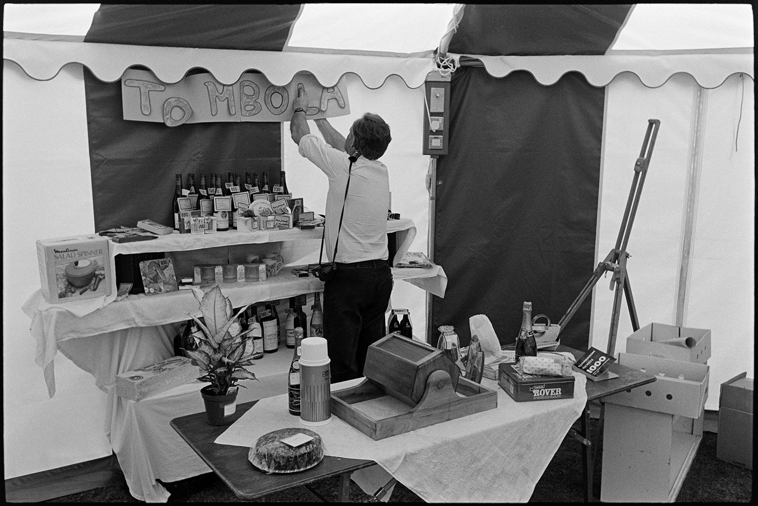 Women decorating marquee tent for hunt ball, flowers, laying tables, tombola, clipping lawn.
[A man setting up a tombola stall in a marquee for the Eggesford Hunt Ball at Colleton Manor, Chulmleigh. Various prizes including bottles of wine, a salad spinner and a cake are visible on the stall. He has a camera over his shoulder.]