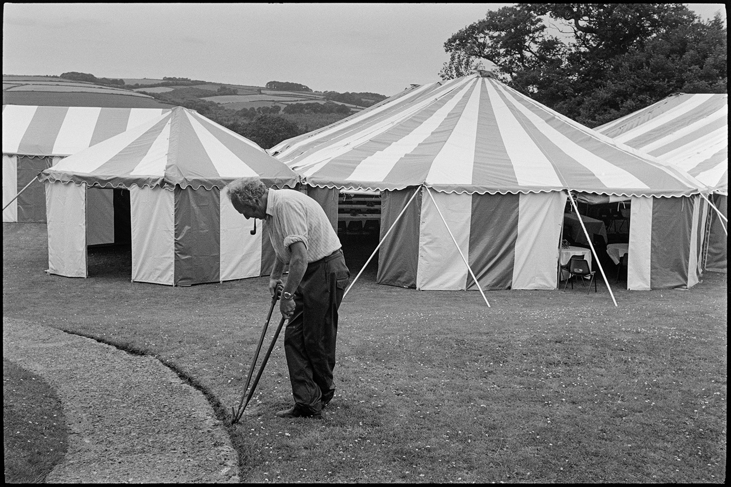 Women decorating marquee tent for hunt ball, flowers, laying tables, tombola, clipping lawn.
[A man clipping the edge of a path outside the marquees set up for the Eggesford Hunt Ball at Colleton Manor, Chulmleigh. He is smoking a pipe.]