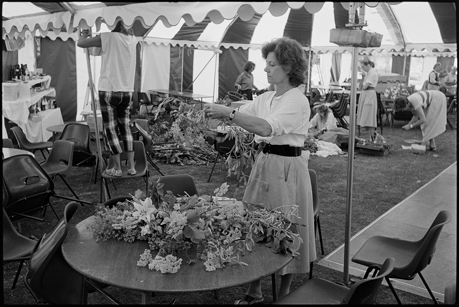 Women decorating marquee tent for hunt ball with flowers.
[Women decorating the inside of a marquee with flowers for the Eggesford Hunt Ball at Colleton Manor, Chulmleigh. A woman is making a flower arrangement at a table in the foreground.]
