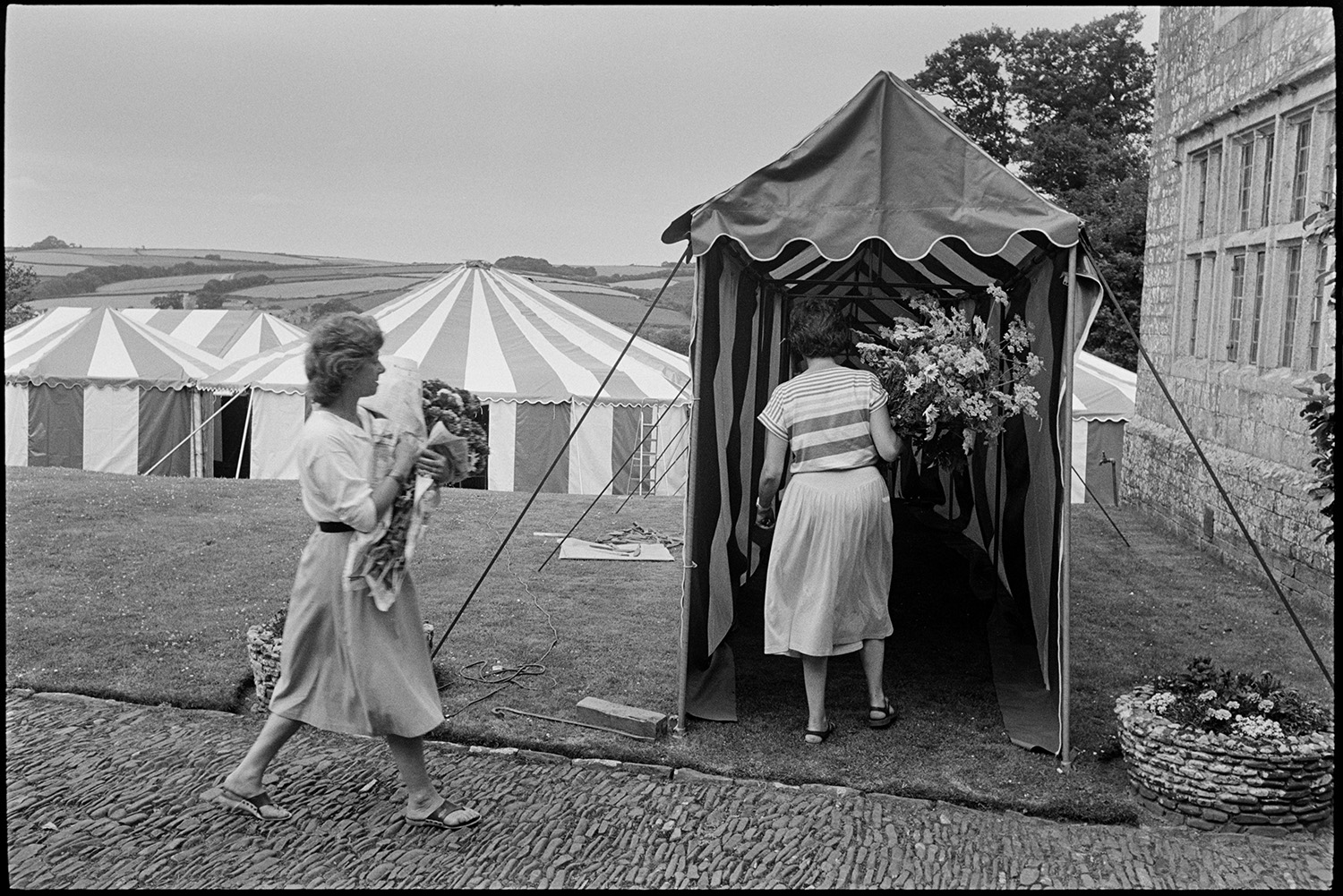 Women decorating marquee tent for hunt ball with flowers.
[Women walking into a marquee entrance with flower arrangements for the Eggesford Hunt Ball at Colleton Manor, Chulmleigh. The marquee entrance is by a cobbled pathway.]