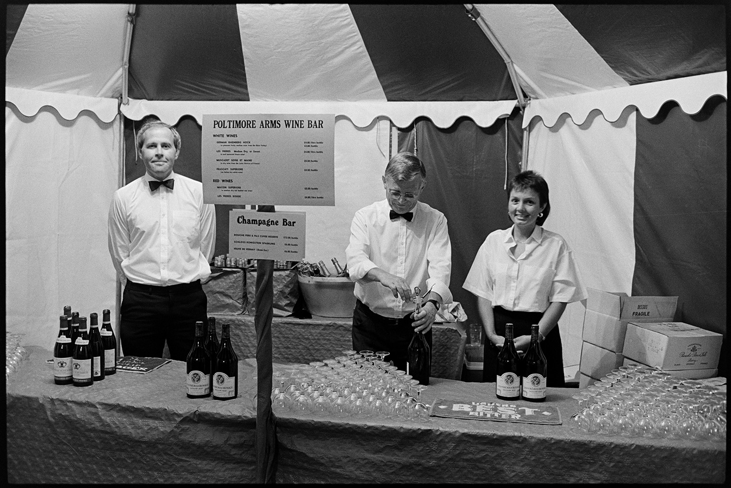 Hunt ball tents marquee, night with candles, people at bar, and arriving, disco lorry.
[Staff standing at the Poltimore Arms Wine Bar in a marquee for the Eggesford Hunt Ball at Colleton Manor, Chulmleigh. Bottles of wine and glasses are laid out on the table in front of the staff.]
