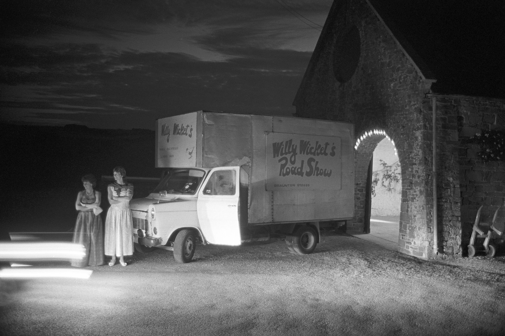 Hunt ball disco lorry with two women arriving in long dresses at night, lit by headlights. 
[Two women in evening dresses stood by a small lorry with the Willy Wicket's Road Show or disco for the hunt ball at Colleton Manor, Chulmleigh, at night. The scene is lit by the headlights of an approaching car.]