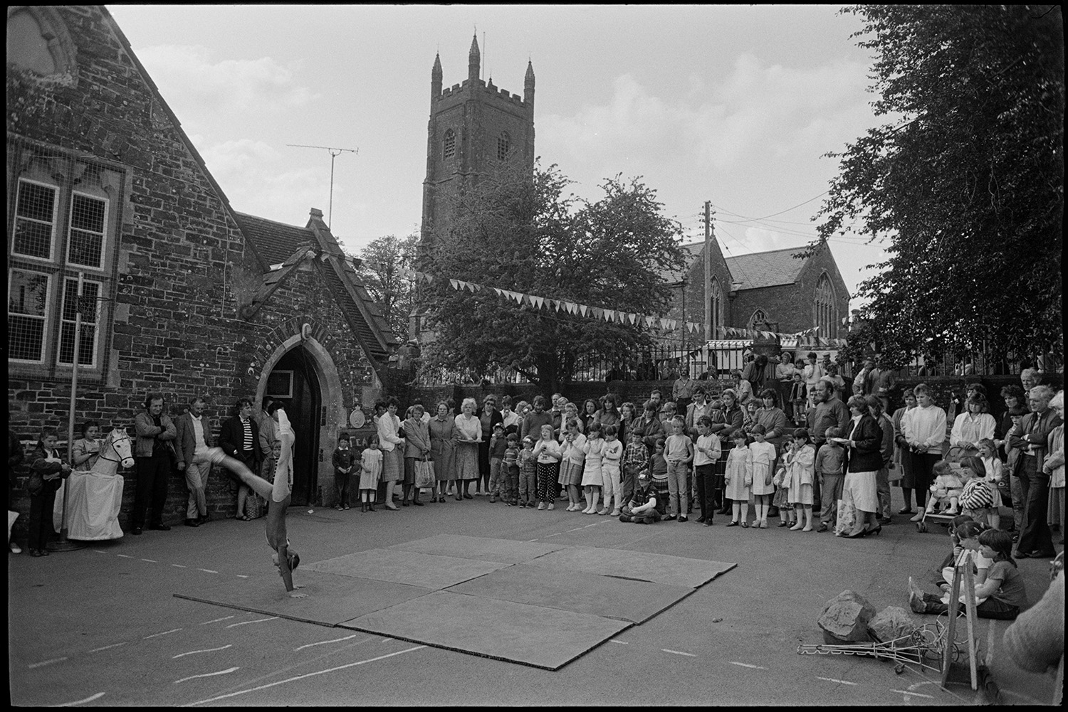 A crowd watching a girl perform a gymnastic display in the playground at Chulmleigh Primary School fete. The playground is decorated with bunting and Chulmleigh Church can be seen in the background.