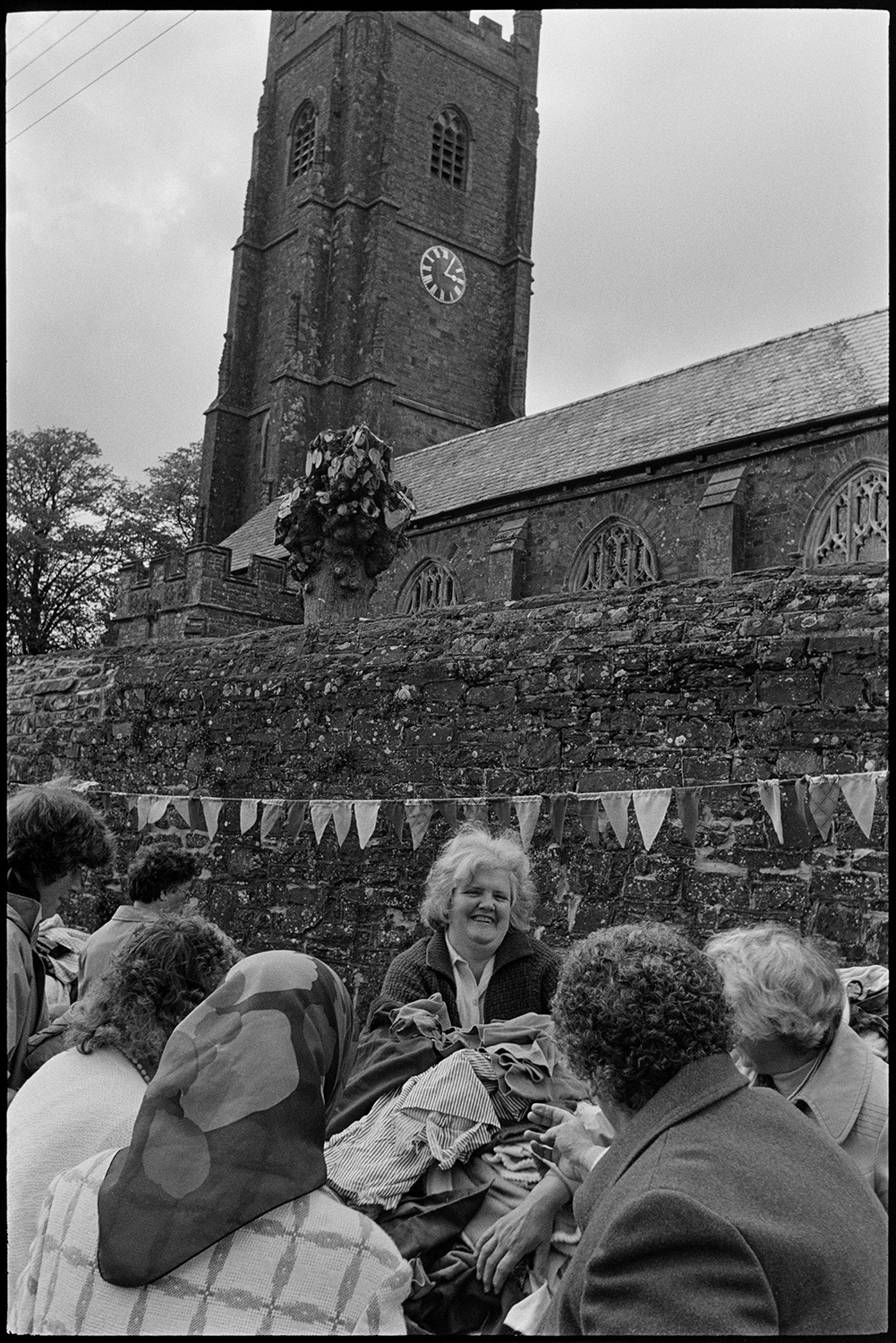 School fete, stalls, gymnastic display in school playground. 
[Women looking at a jumble stall  at Chulmleigh Primary School fete in the playground. The wall behind the stall is decorated with bunting and Chulmleigh Church is visible in the background.]