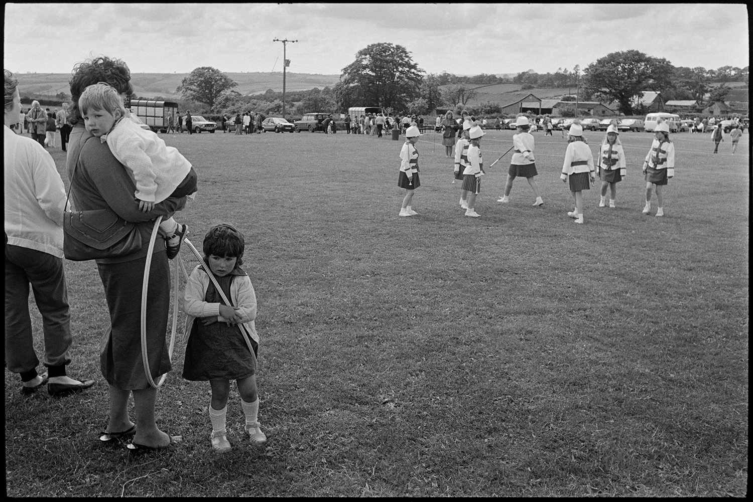 School fete, drum majorettes competition, early tractor display, mothers and children.
[A woman with two young children holding hula-hoops, watching majorettes getting ready to perform at the Chulmleigh School Fete, in a field. Parked cars and trailers can be seen in the background.]